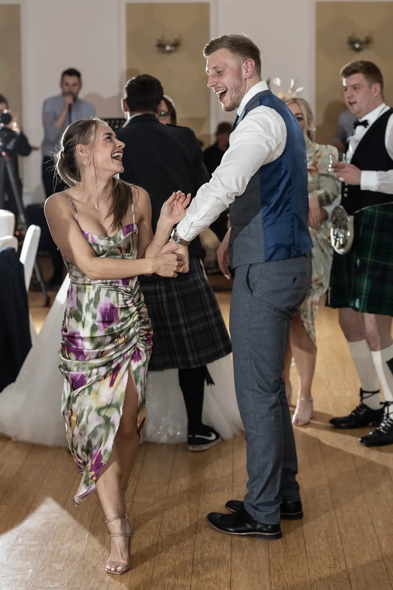 A smiling woman in a floral dress and a man in a grey suit with a blue waistcoat dance together joyfully at a wedding reception. Other guests, including a man in a kilt, stand and mingle in the background on a wooden floor.