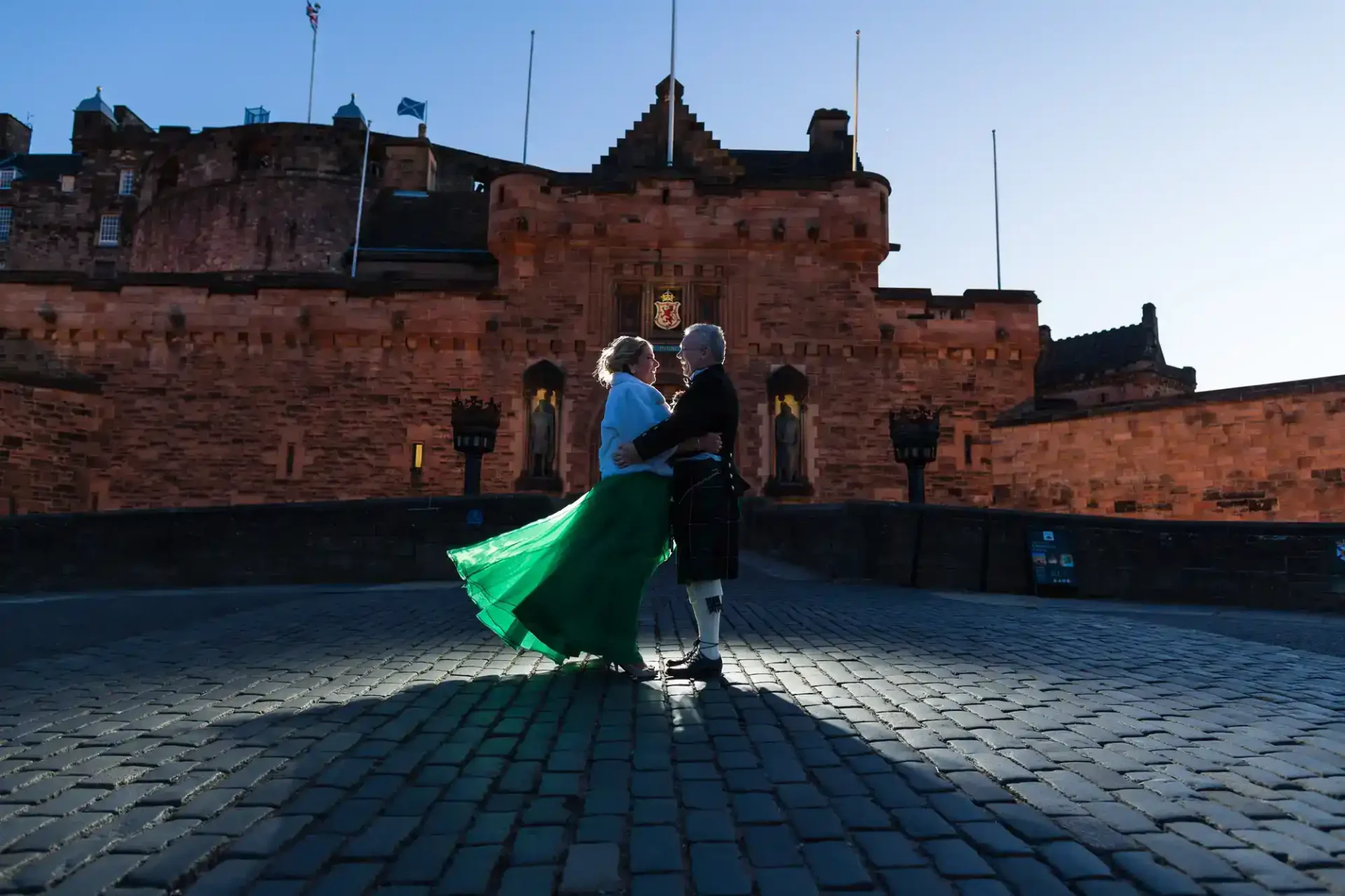 An older couple dressed in formal attire, including a green skirt and a kilt, stand together on a cobblestone path in front of historic Edinburgh Castle at dusk.