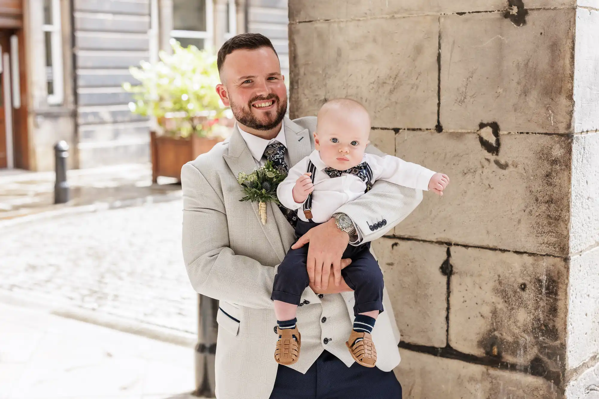 A smiling man in a light suit holds a baby in his arms. The baby wears navy trousers, braces, and a bow tie. They stand outside by a stone wall, with sunlight and a building with plants in the background.