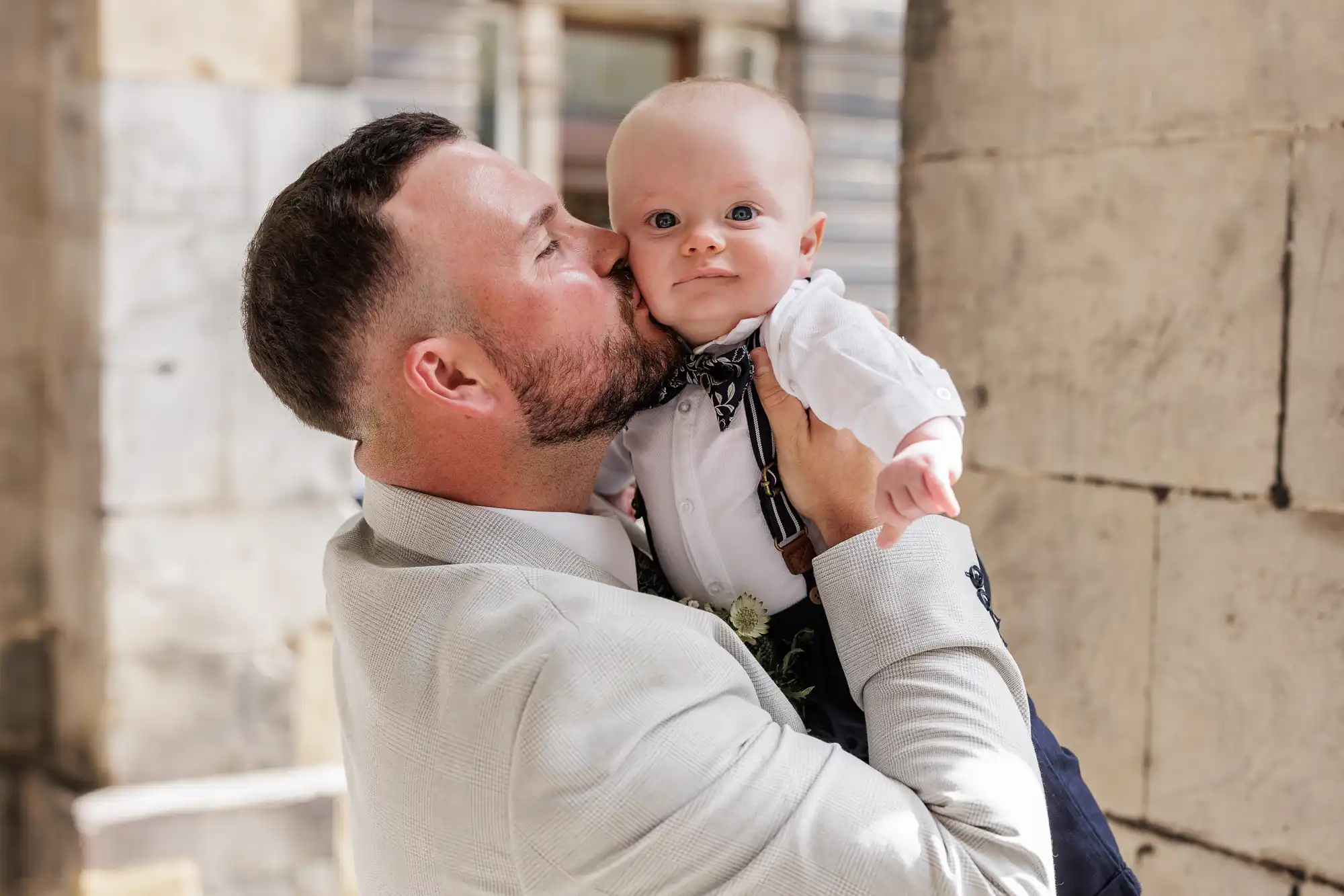 A man in a light suit lifts and kisses a baby on the cheek. The baby, dressed in a white shirt with dark braces and a bow tie, looks at the camera. They are standing next to a stone wall in a sunlit outdoor setting.