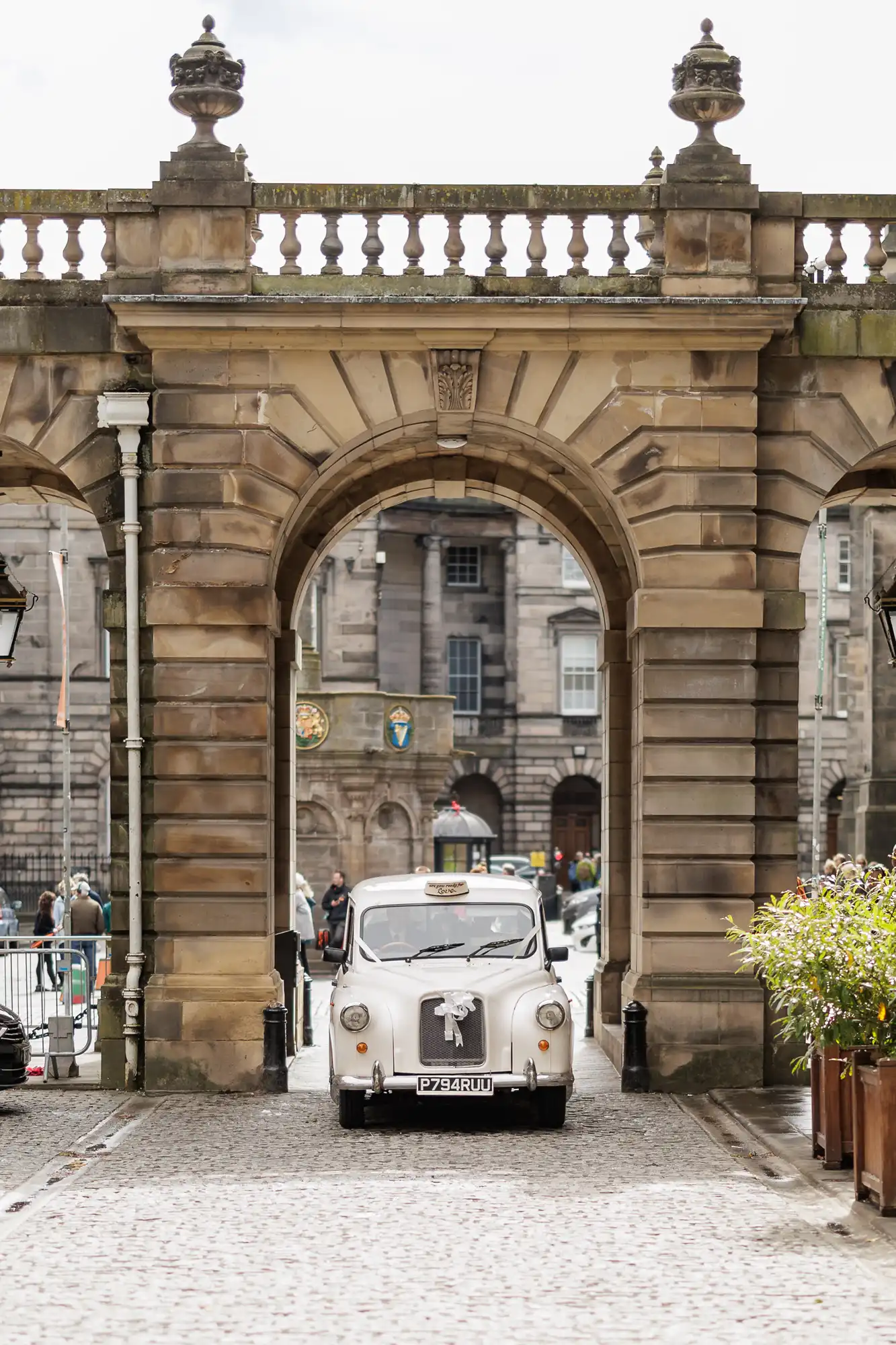 A classic white taxi decorated with white ribbons is parked under a large stone archway in a historic, cobbled courtyard. The background features grand stone buildings, iron gates, and people walking nearby.