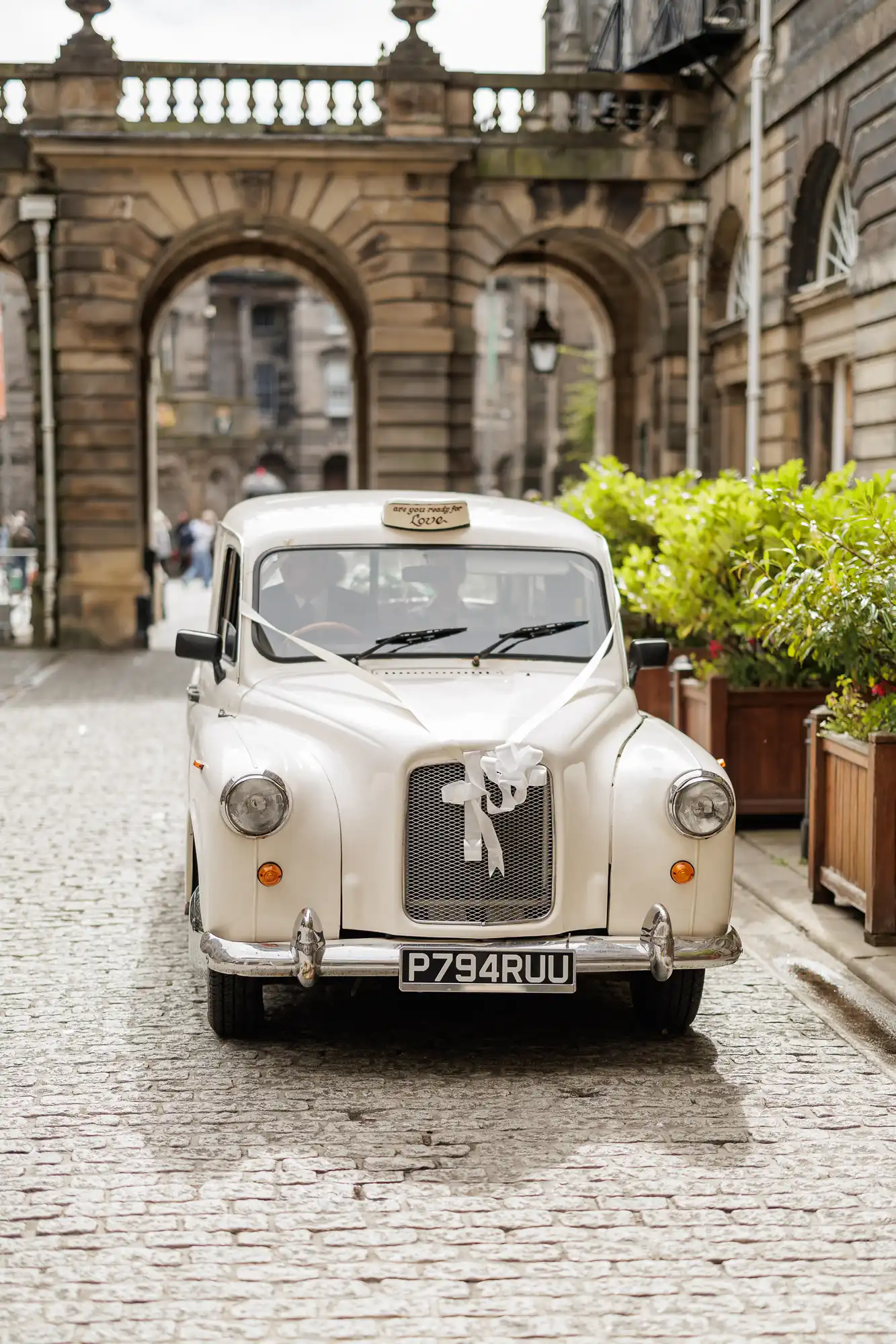 A classic white taxi decorated with a white ribbon and a "Just Married" sign is parked on a cobbled street, surrounded by stone buildings and planters with green plants. An arched entrance appears in the background.