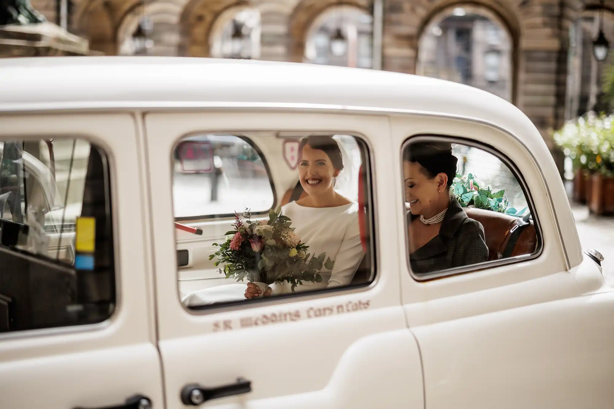Two women dressed for a wedding sit in the back seat of a white vintage car. One holds a bouquet and smiles, wearing a white dress, while the other, in dark attire and pearls, sits beside her. Sunlight streams through the car window, revealing stone arches outside.