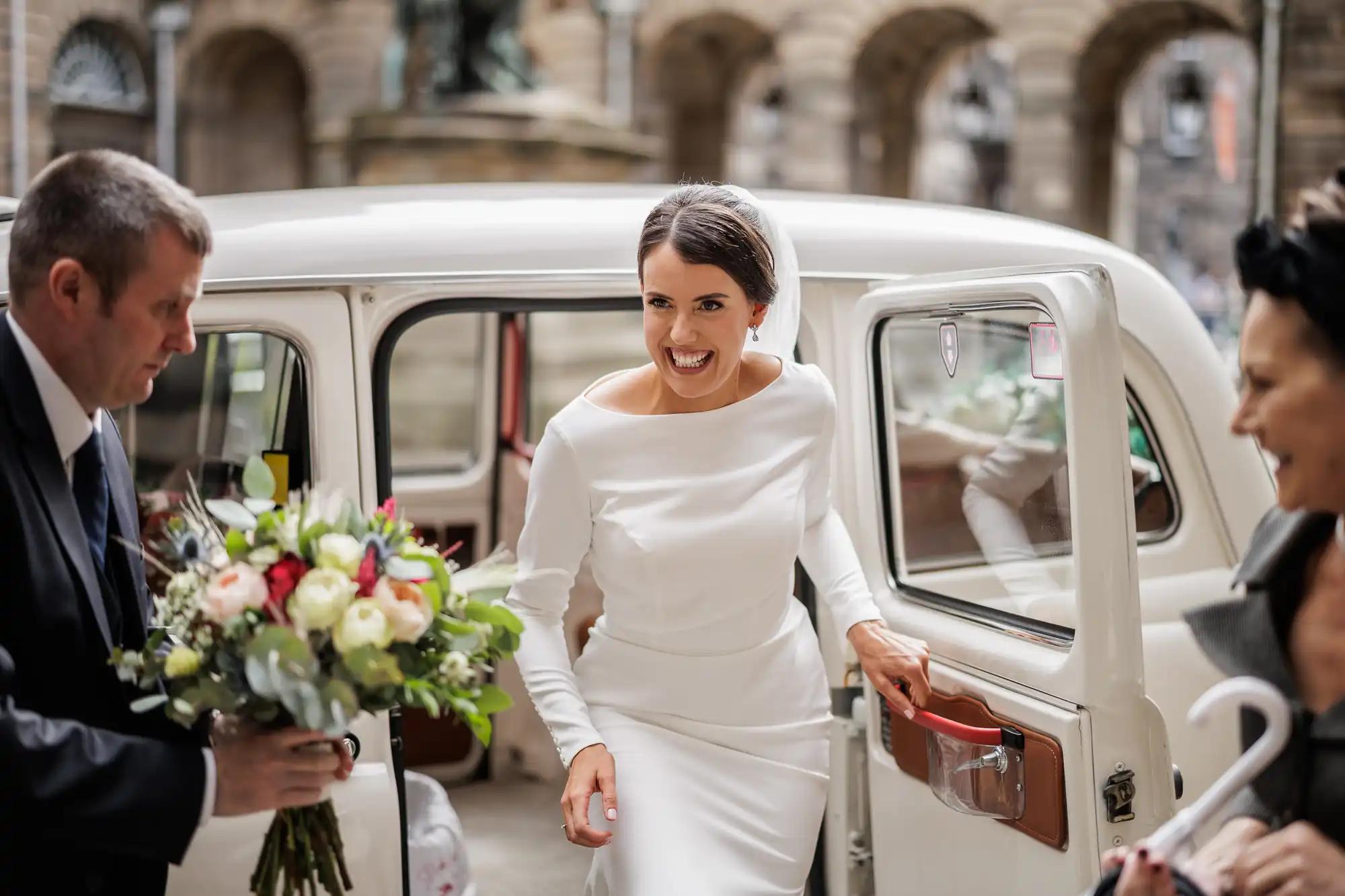 A smiling bride in a white dress steps out of a classic white car. A man in a suit holds a bunch of flowers for her, while another woman, slightly blurred, stands nearby. The background features old stone buildings with arches.