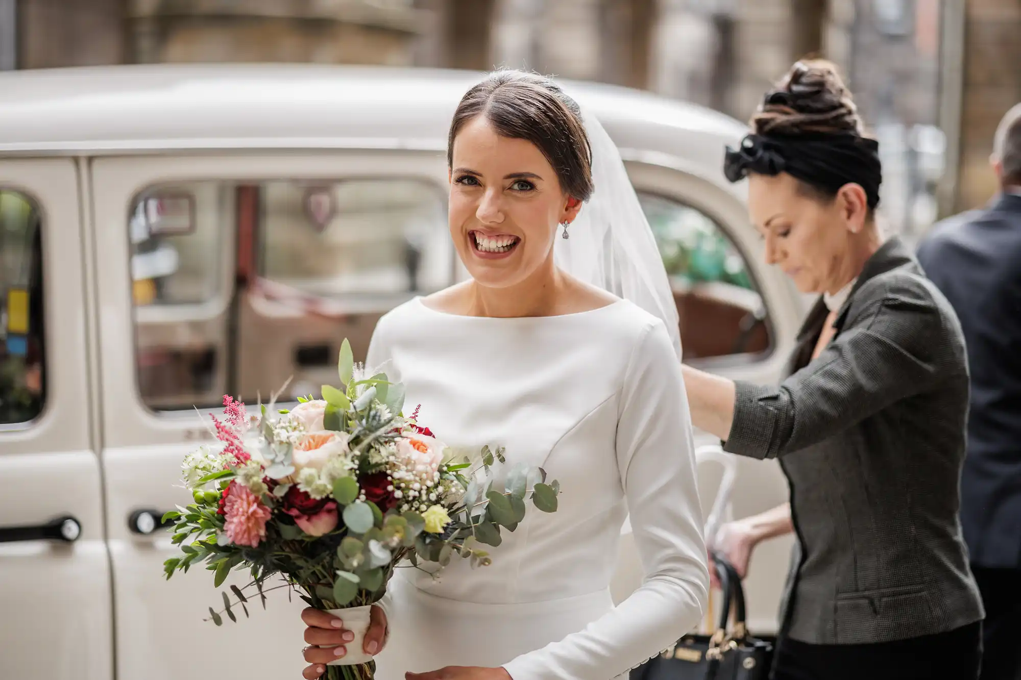 A smiling bride in a white dress and veil holds a colourful bouquet of flowers while standing outside next to a white vintage car. In the background, another woman adjusts the car door. The setting appears urban with stone buildings visible.