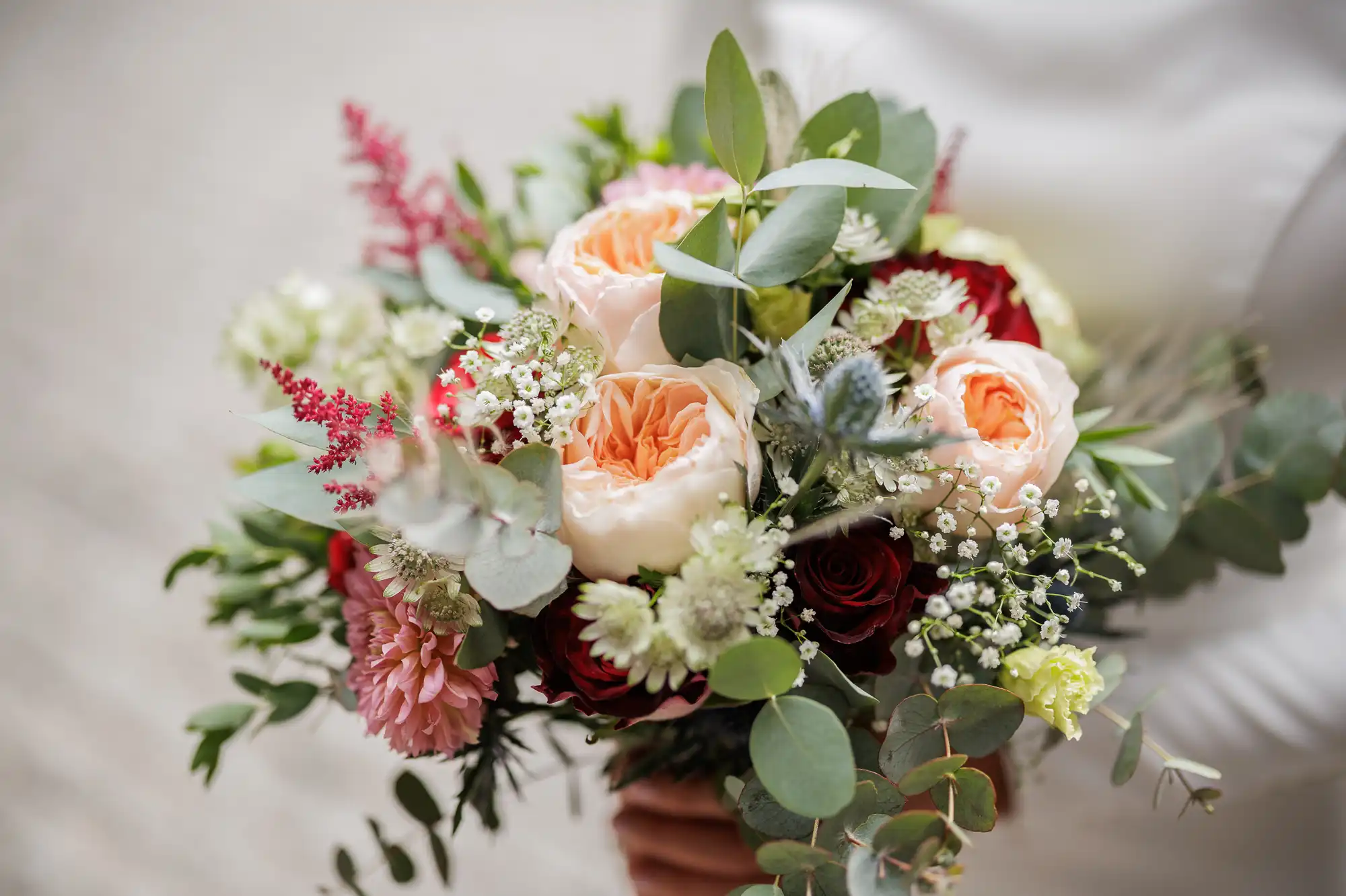 A close-up of a vibrant bouquet featuring peach roses, red roses, white baby's breath, pink flowers, green eucalyptus leaves, and assorted greenery, held by a person in white attire with a soft blurred background.