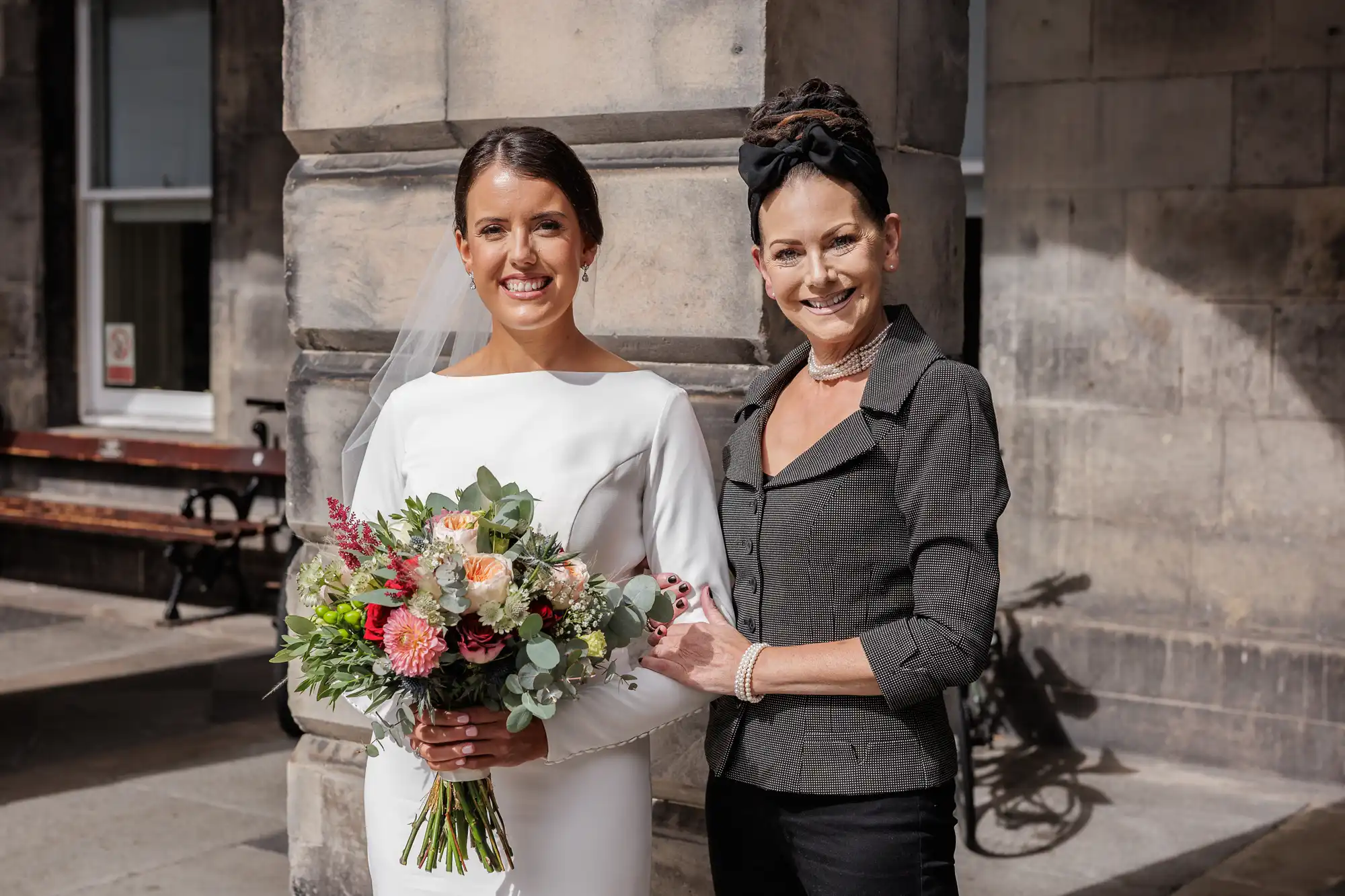 A bride in a long-sleeved white dress holds a bouquet of colourful flowers and stands beside a smiling woman in a dark jacket and pearl necklace, posing outdoors in front of a stone building under bright sunlight.
