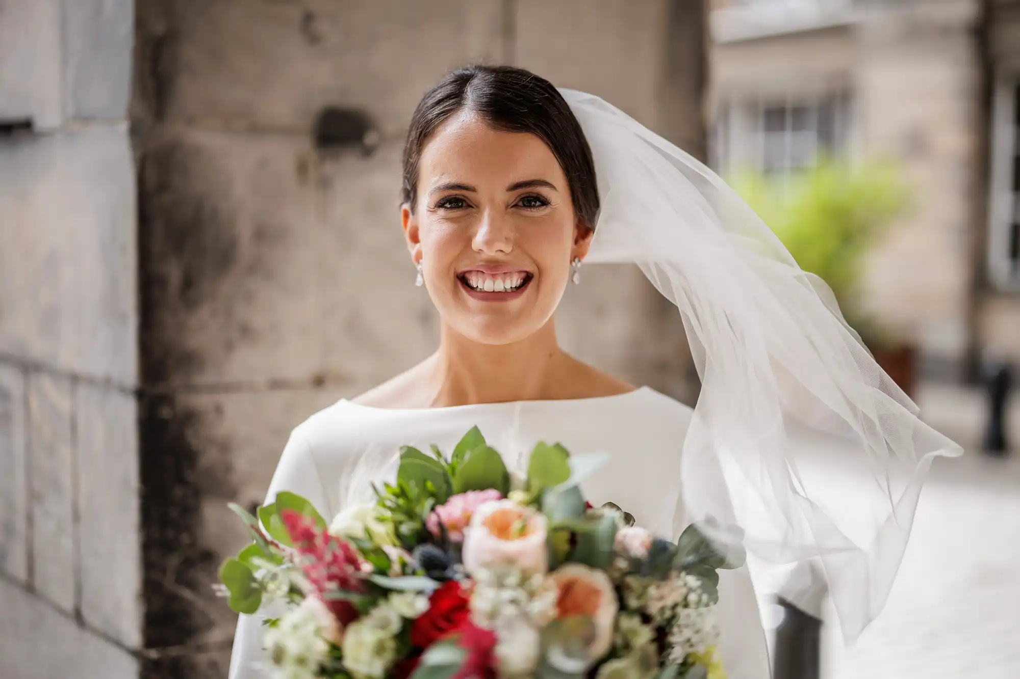 A smiling bride stands outdoors, holding a colourful bouquet of flowers with pink, white, and green tones. She wears a white dress and a veil that flows to the side. The background is softly blurred with stone walls and greenery visible.
