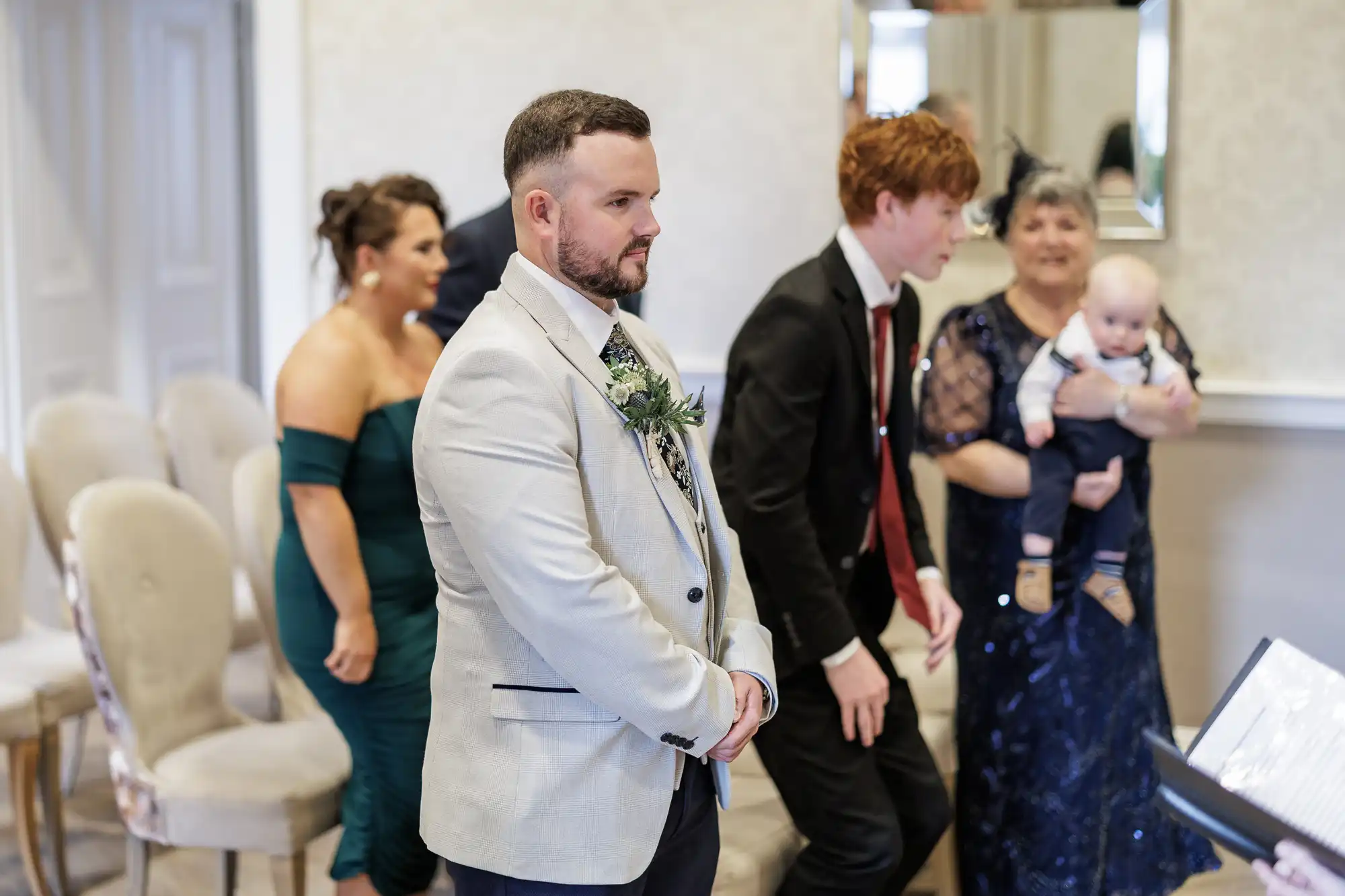 A man in a cream suit with a buttonhole stands at the front of a room. Behind him, a woman in a green dress, a man in a black suit, an older woman in a navy dress holding a baby, and another person are present. Some are standing; chairs line the room.