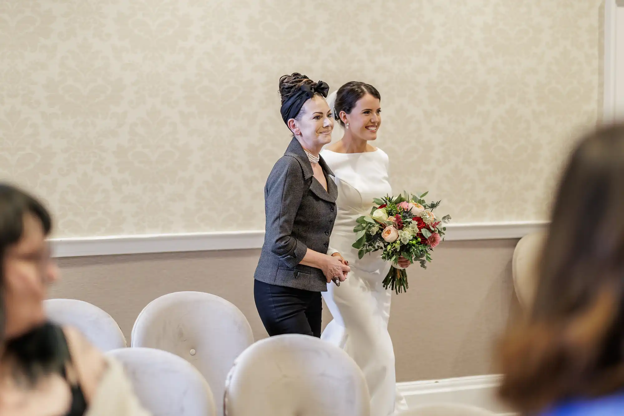 A smiling bride in a white dress, holding a bouquet of flowers, walks down the aisle beside a woman in a grey jacket. Both women look happy. There are empty, light-coloured chairs and a blurred guest in the foreground. The setting is indoors with patterned walls.