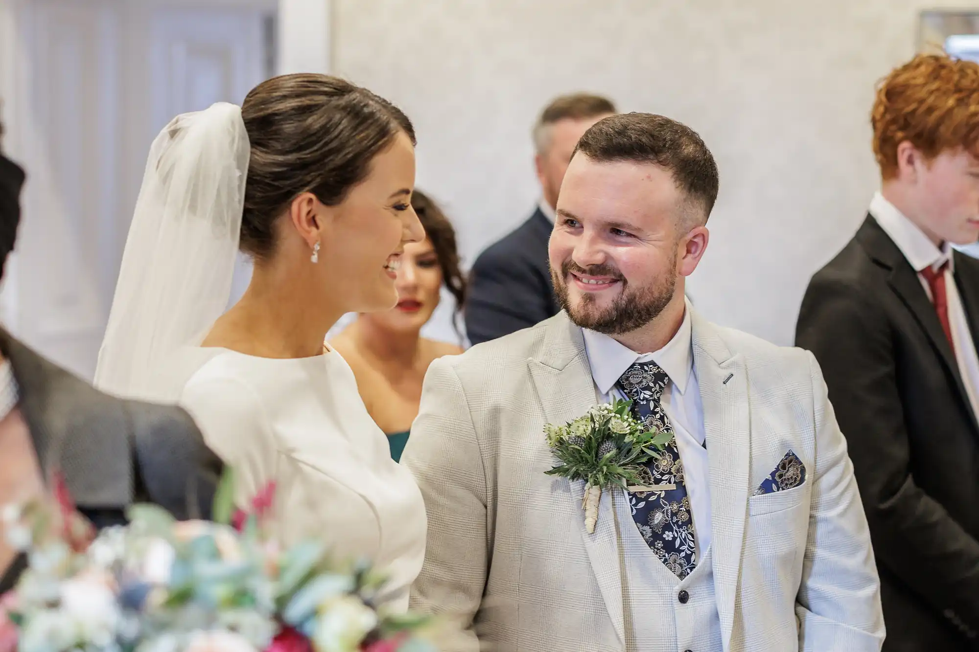 A bride in a white dress and veil smiles at a groom in a light suit with a floral buttonhole. They stand close, facing each other. Guests in formal attire are blurred in the background, suggesting a joyful wedding ceremony indoors.