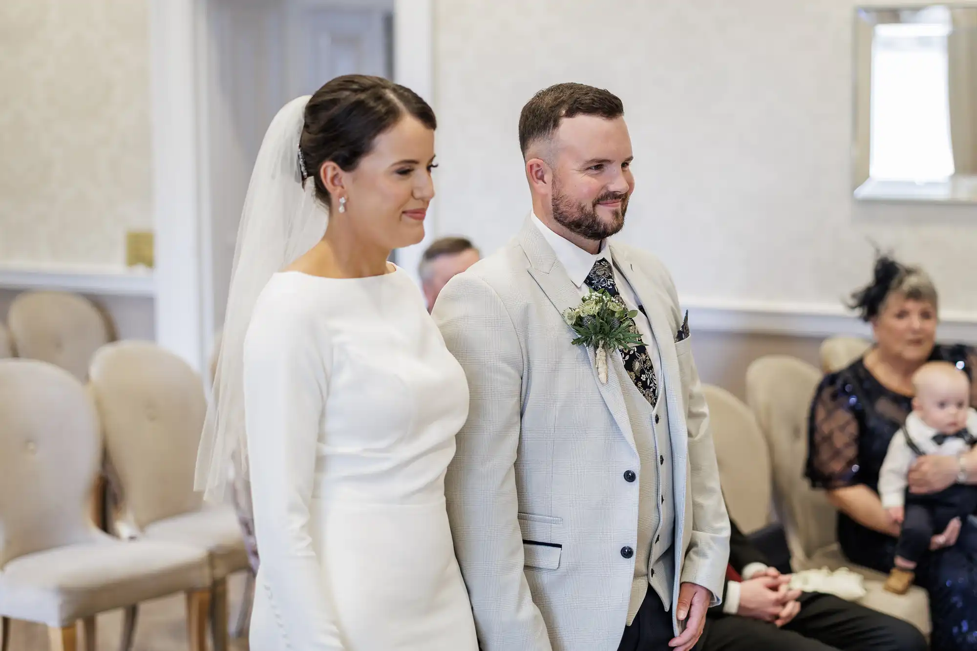 A bride in a long-sleeved white dress and veil stands beside a groom in a light suit with a floral buttonhole. They smile during a wedding ceremony, with guests seated behind them, including a woman holding a baby. The room is softly lit and elegant.