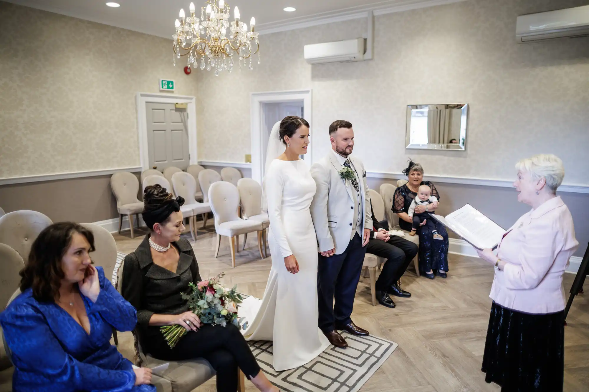 A bride in a white dress and a groom in a light suit stand holding hands in a small, elegant room with cream chairs. A celebrant reads from a booklet; guests, including women and a baby, are seated. A chandelier hangs above. The mood is intimate and joyful.