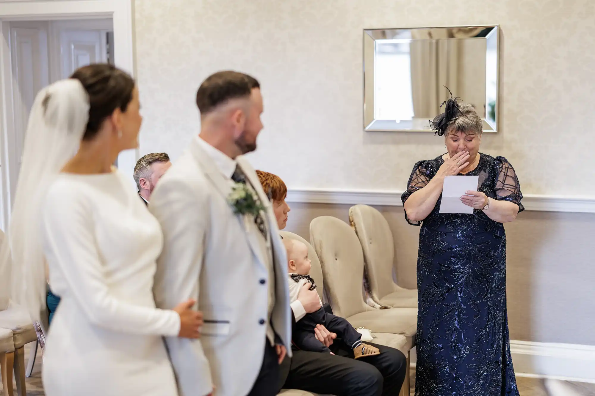 A woman in a navy dress stands reading from a paper, appearing emotional with her hand covering her mouth. A bride in a white dress and veil stands arm-in-arm with a groom in a light suit, both watching her. Seated guests, including a child, listen attentively.