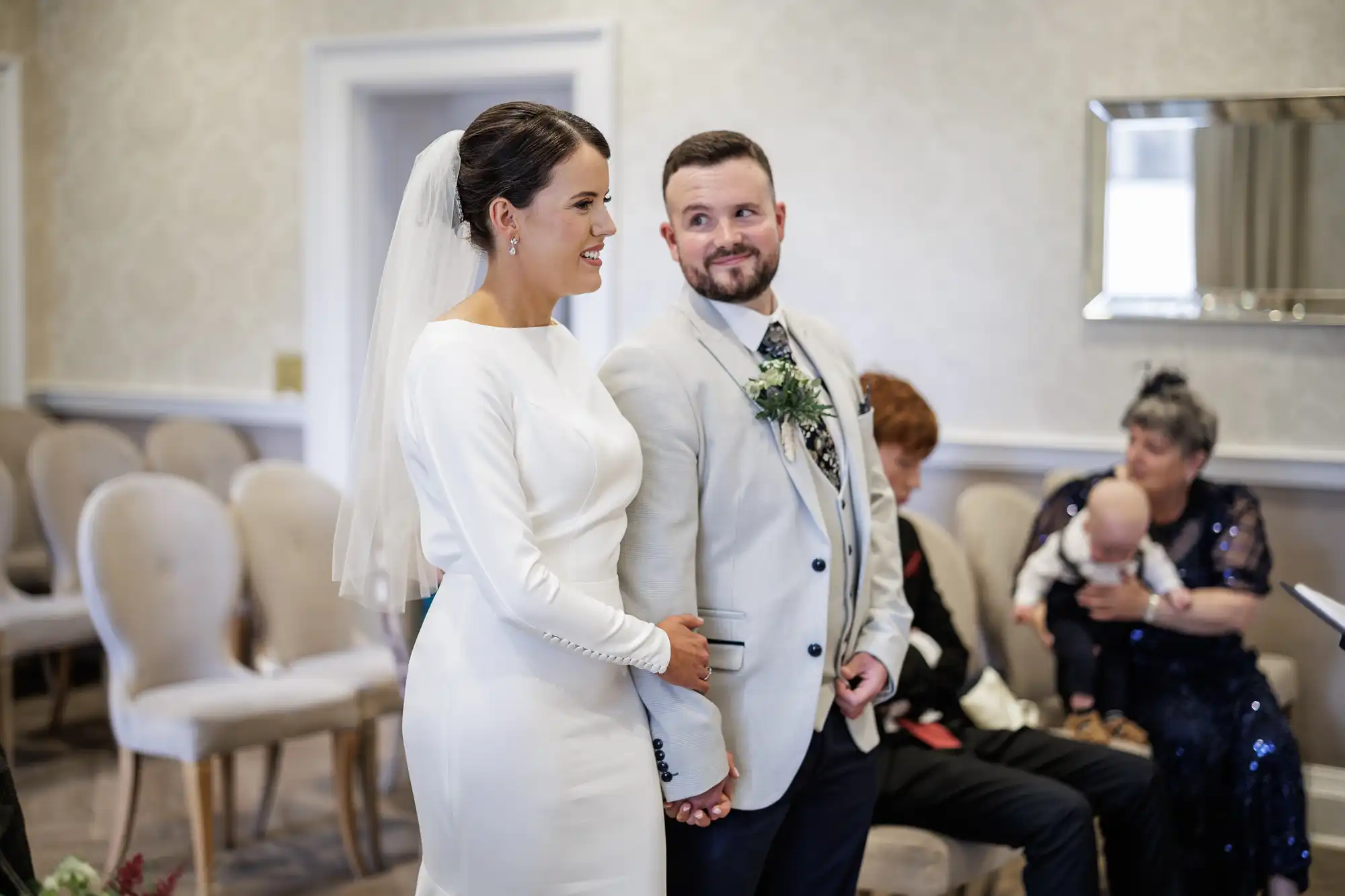 A bride in a white dress and veil holds hands with a smiling groom in a light suit and tie. They stand in a small room with cream chairs. Guests, including an older woman holding a baby, are seated behind them. The mood is joyful and intimate.
