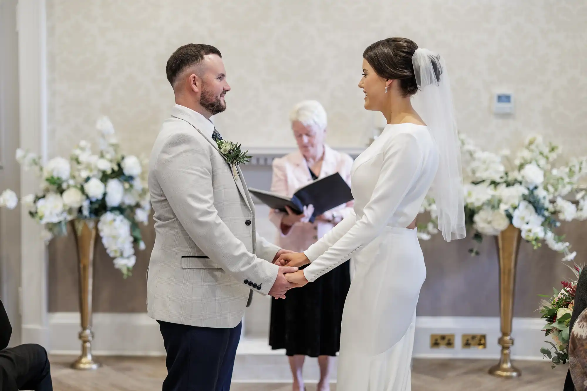 A bride and groom stand facing each other, holding hands and smiling, during their wedding ceremony. The bride wears a white dress and veil; the groom is in a light suit. An officiant stands behind them holding a book. Floral arrangements decorate the background.