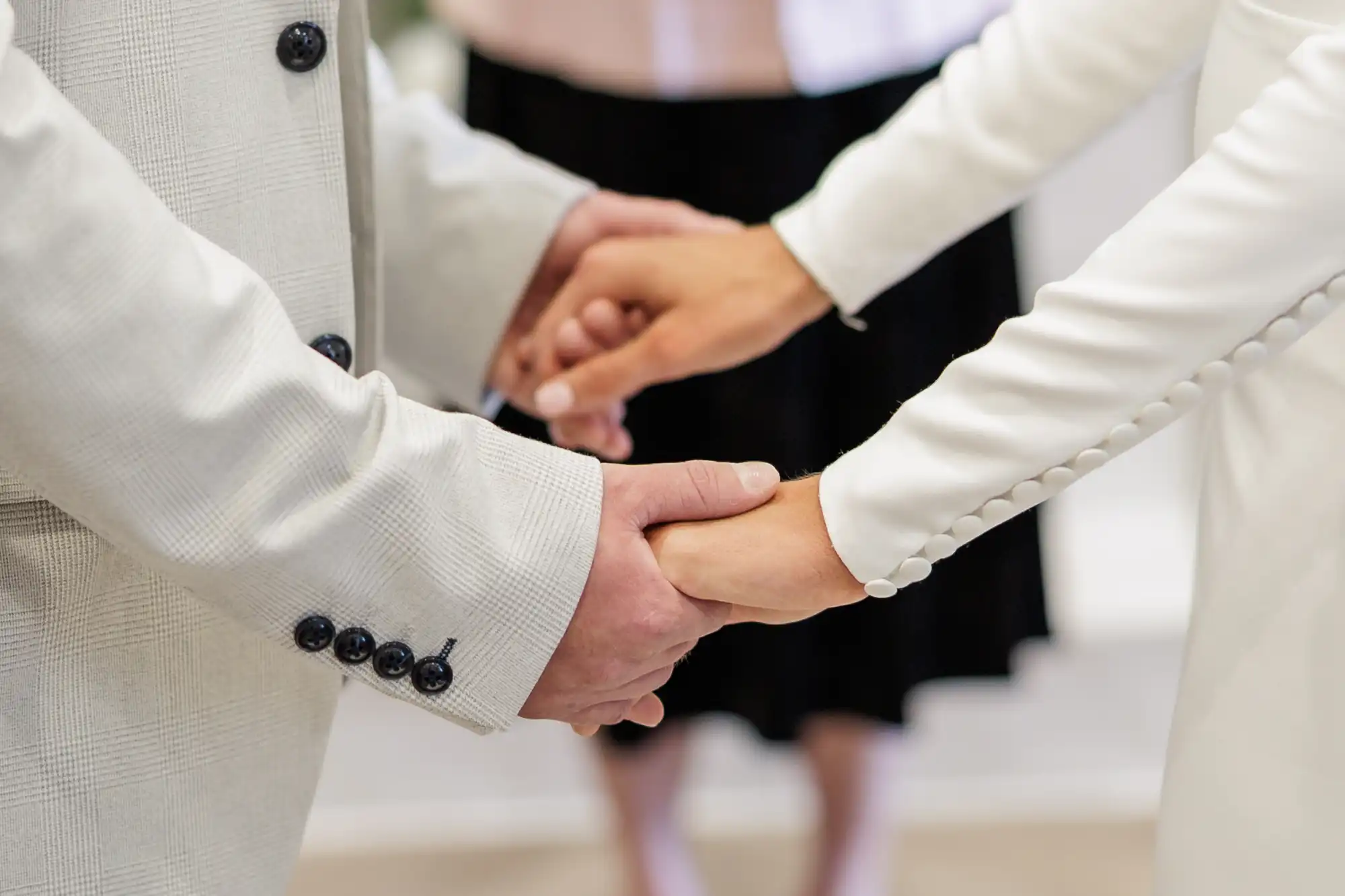 Close-up of two people in light-coloured formal attire holding hands, possibly during a wedding ceremony. A person in the background is out of focus, wearing a light top and dark skirt. The scene suggests a moment of commitment or celebration.