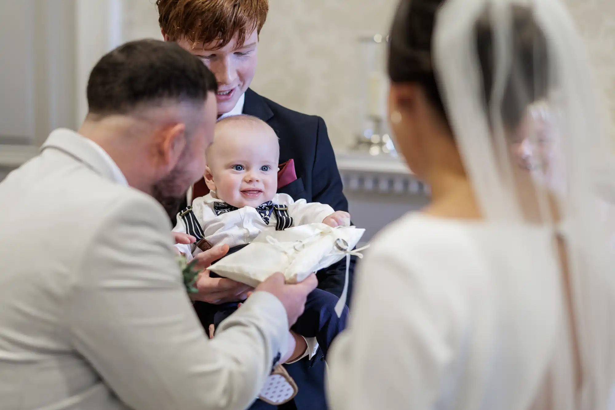 A smiling baby in a bow tie and striped braces is held by a man in a suit during a wedding ceremony. Two adults, one in a white suit and one in a wedding dress with a veil, face the baby, who is reaching for a ring cushion. The scene is joyful and formal.