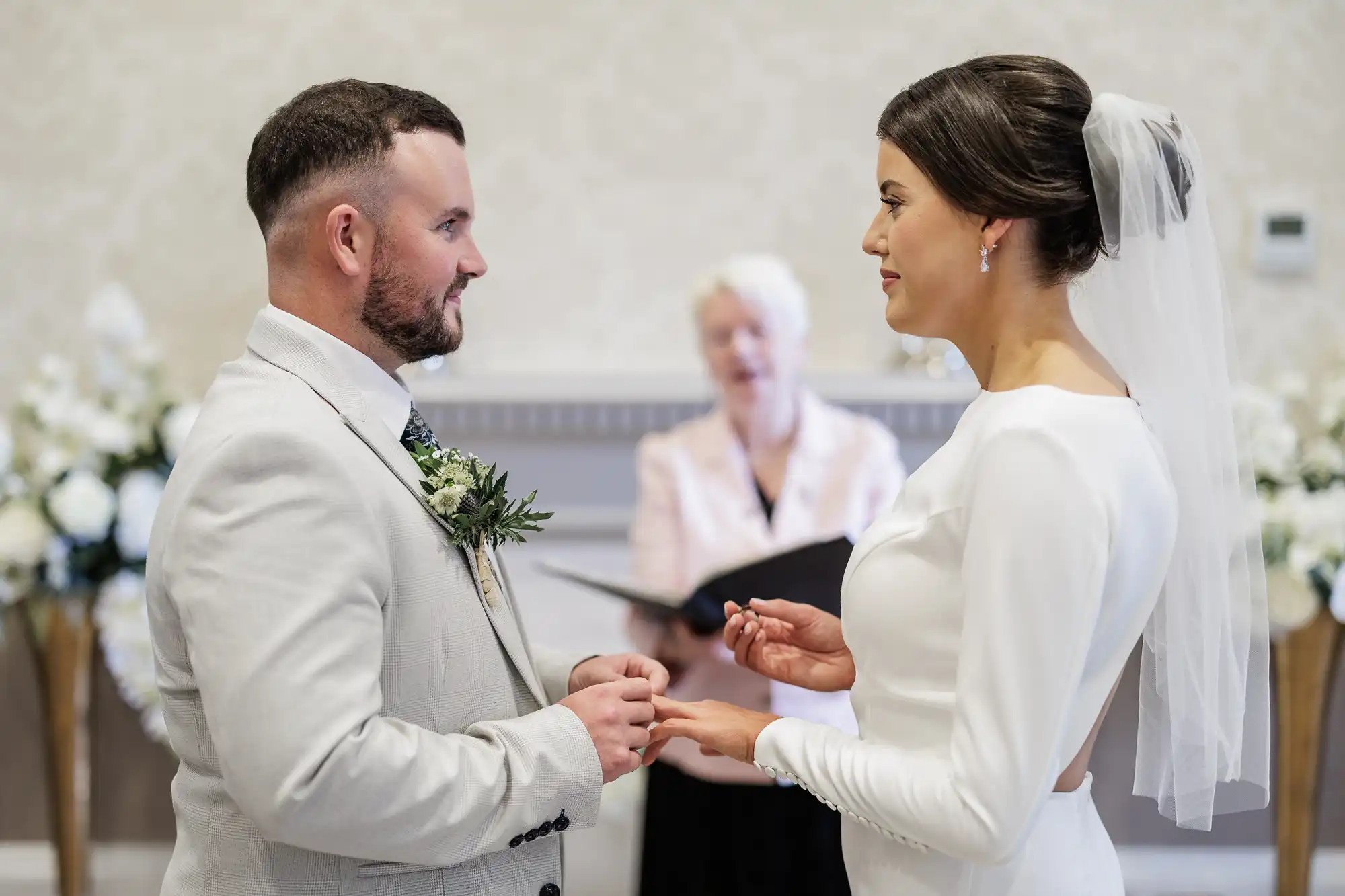A groom in a light suit places a ring on the bride’s finger as they face each other, smiling. The bride wears a white dress and veil. An officiant stands behind them holding a book. White flowers and elegant décor are in the background.