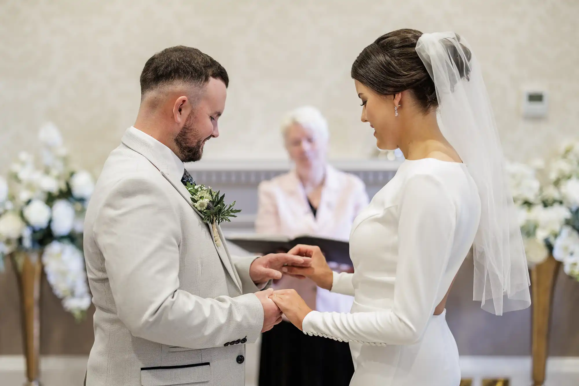 A bride and groom exchange rings during their wedding ceremony. The bride wears a white dress and veil, and the groom wears a light suit with a buttonhole. An officiant stands behind them, and white flowers decorate the background.