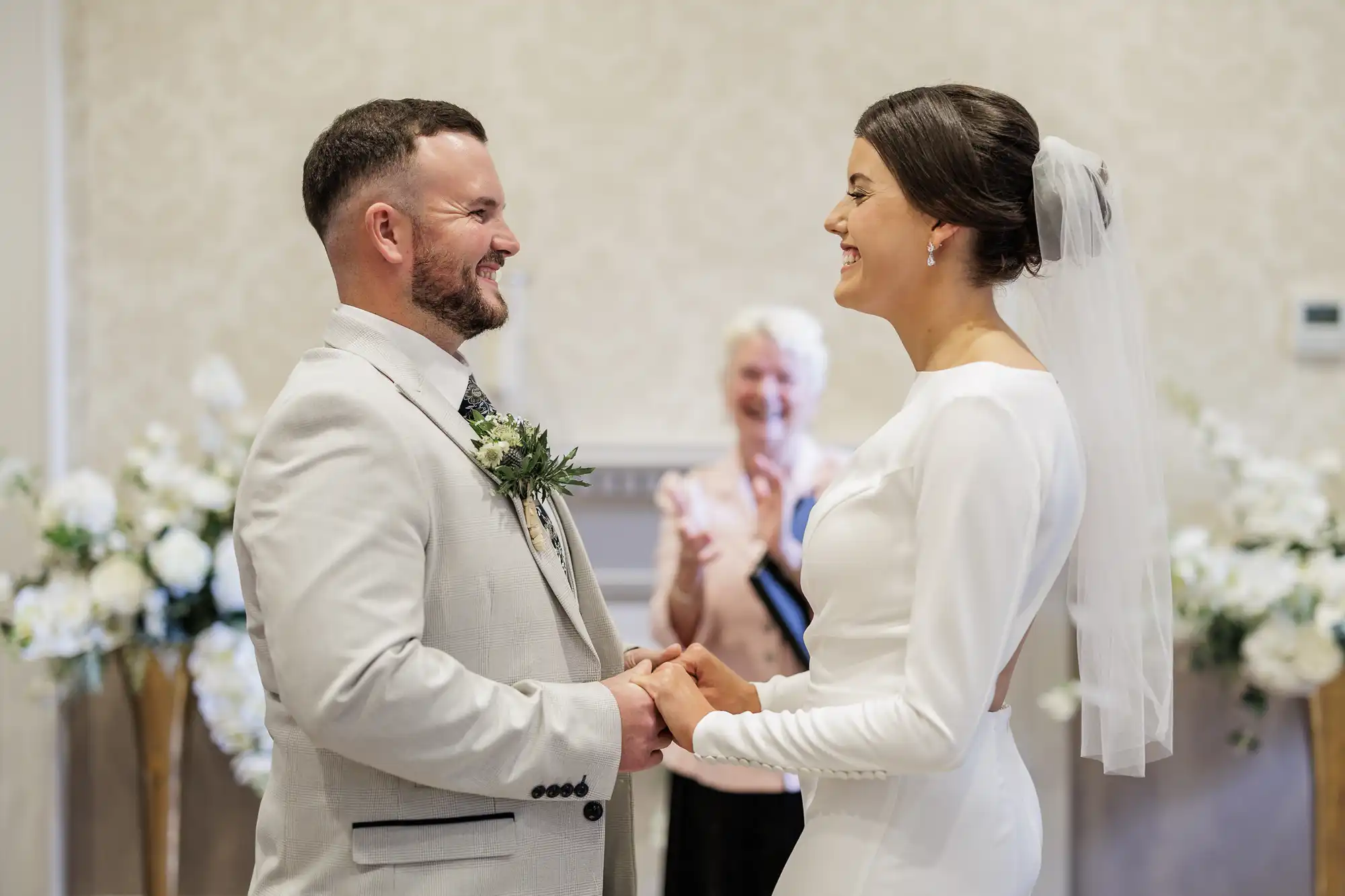 A bride and groom stand facing each other, smiling and holding hands during their wedding ceremony. The bride wears a white dress and veil; the groom is in a light suit with a buttonhole. An officiant smiles and claps in the background amid white floral arrangements.