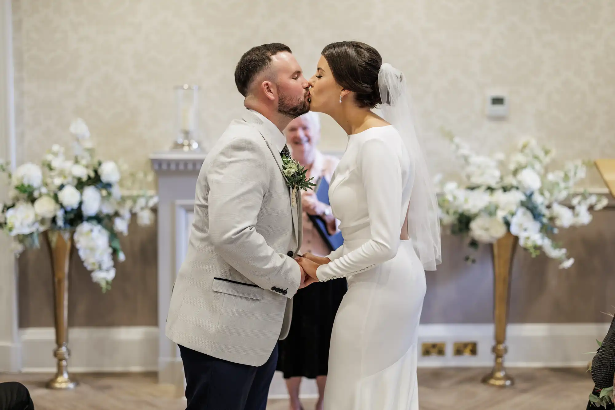 A bride and groom kiss whilst holding hands at their wedding ceremony. The groom wears a light grey suit; the bride wears a long-sleeved white dress and veil. They stand indoors with white floral arrangements and a smiling officiant in the background.