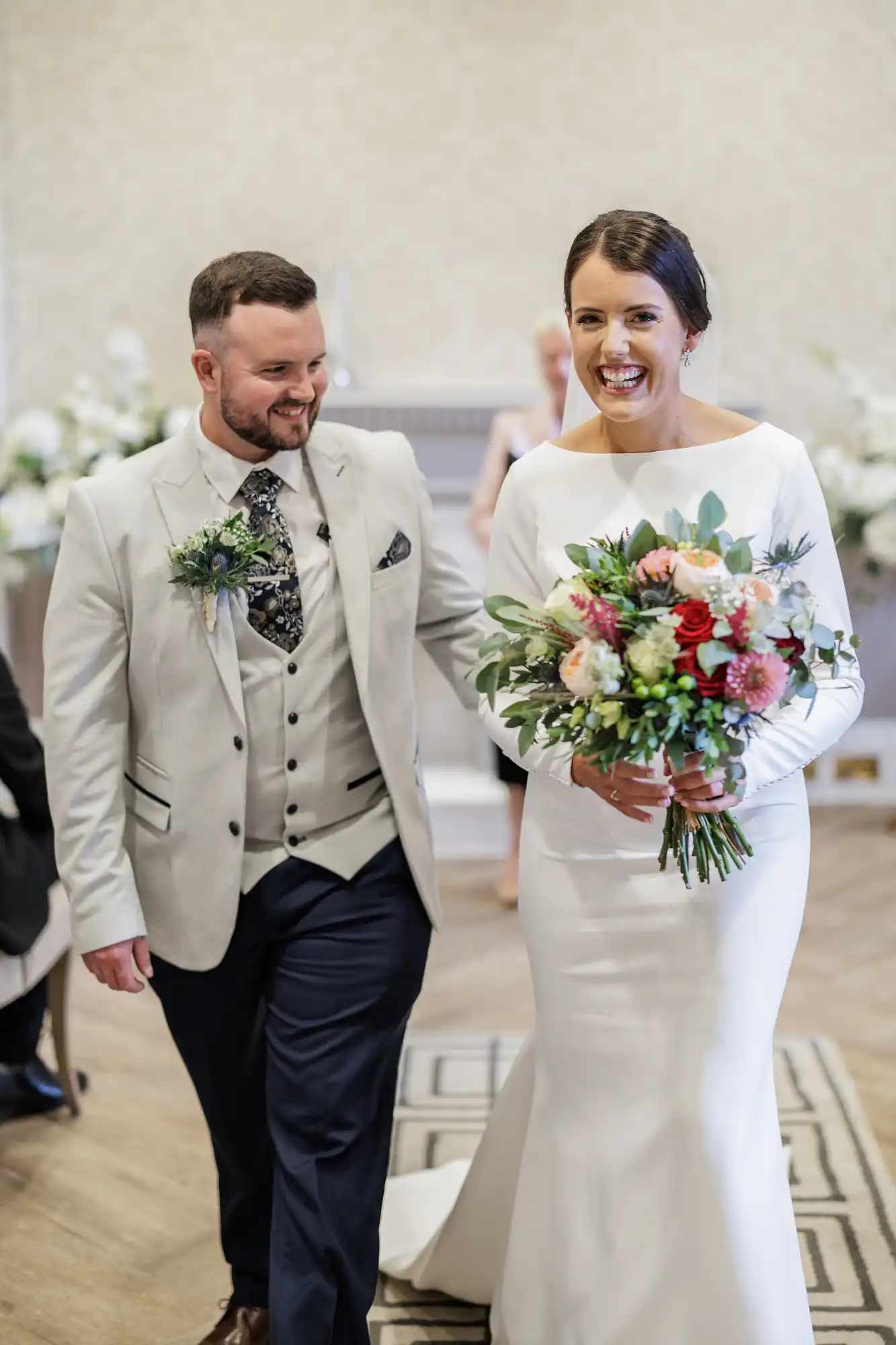 A smiling bride in a long-sleeved white dress holds a colourful bouquet as she walks beside a bearded groom in a light grey jacket, patterned waistcoat, and tie. Both look happy, and floral arrangements decorate the elegant indoor setting.