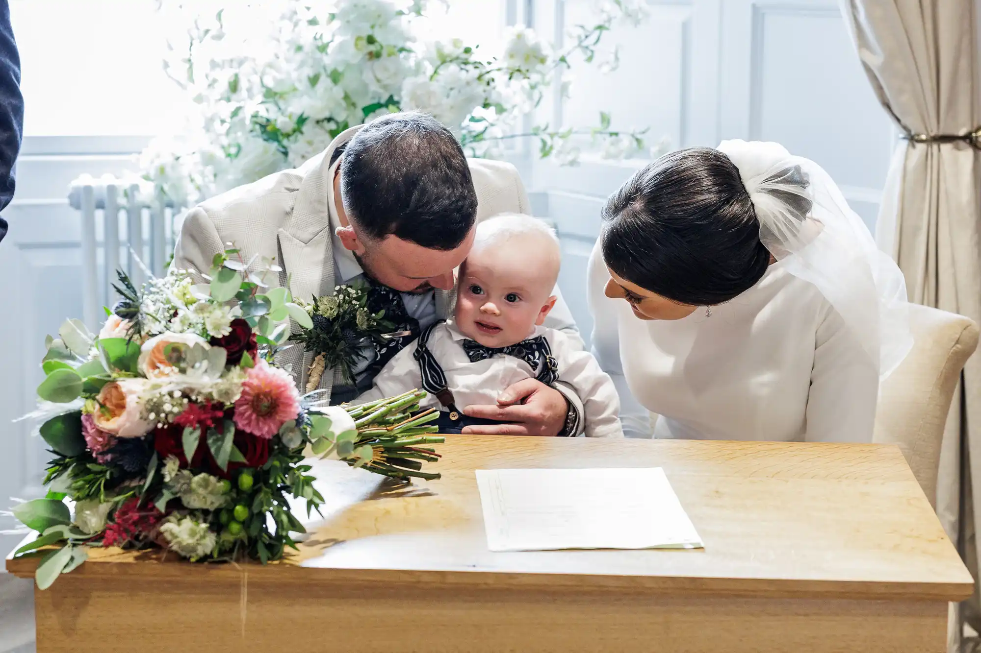 A bride and groom sit closely, holding a baby dressed in a bow tie and braces. The groom kisses the baby's head whilst the bride leans in, smiling. A bouquet of flowers and a document rest on the wooden table in front of them.