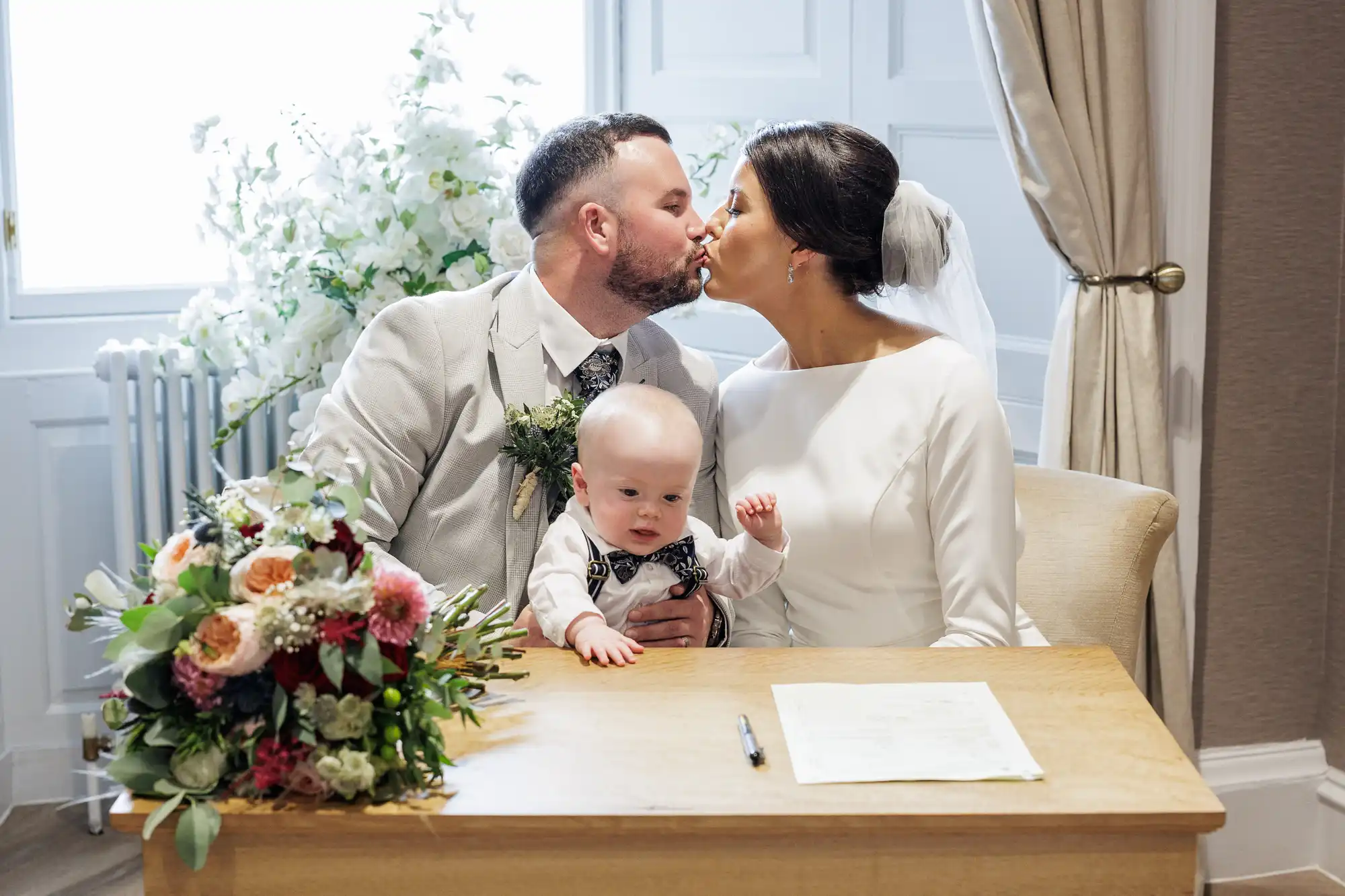 A bride and groom kiss while seated at a table with a baby in the groom’s lap. The bride wears a white dress and veil, and the groom wears a light suit. A bouquet, document, and pen are on the table, with flowers and curtains in the background.