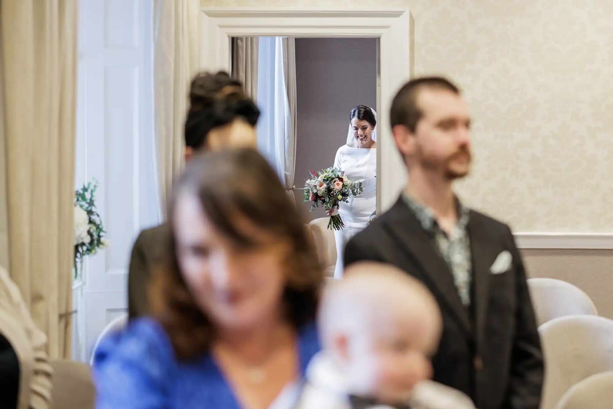 A bride in a white dress holding a bouquet is framed by a doorway in the background, smiling as she enters. In the blurred foreground, seated guests—including a man, a woman in blue, and a baby—look towards her entrance.