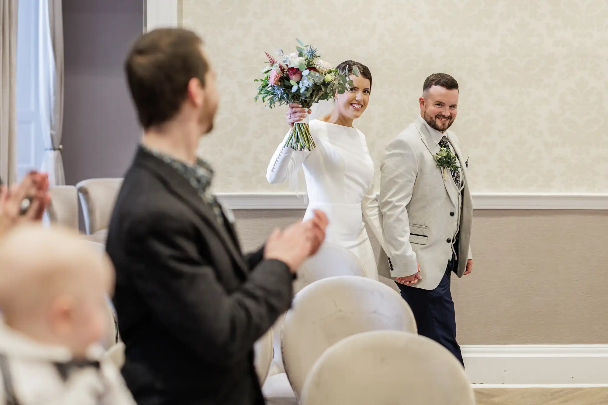 A smiling bride in a white dress holds a bouquet up and walks hand in hand with the groom, who wears a light suit. Guests, including a man in focus and a baby, clap and watch them enter a softly lit, elegant room with cream-coloured walls.