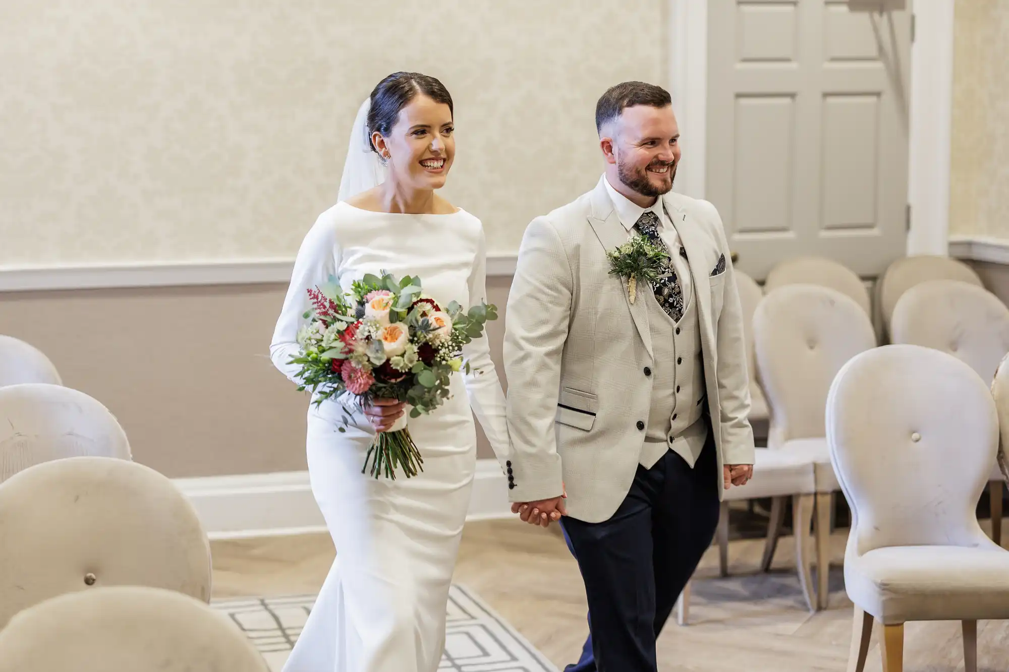 A bride in a white dress holds a bouquet and walks hand-in-hand with a smiling groom in a light-coloured suit. They walk down an aisle lined with empty beige chairs in an elegant, softly lit room.
