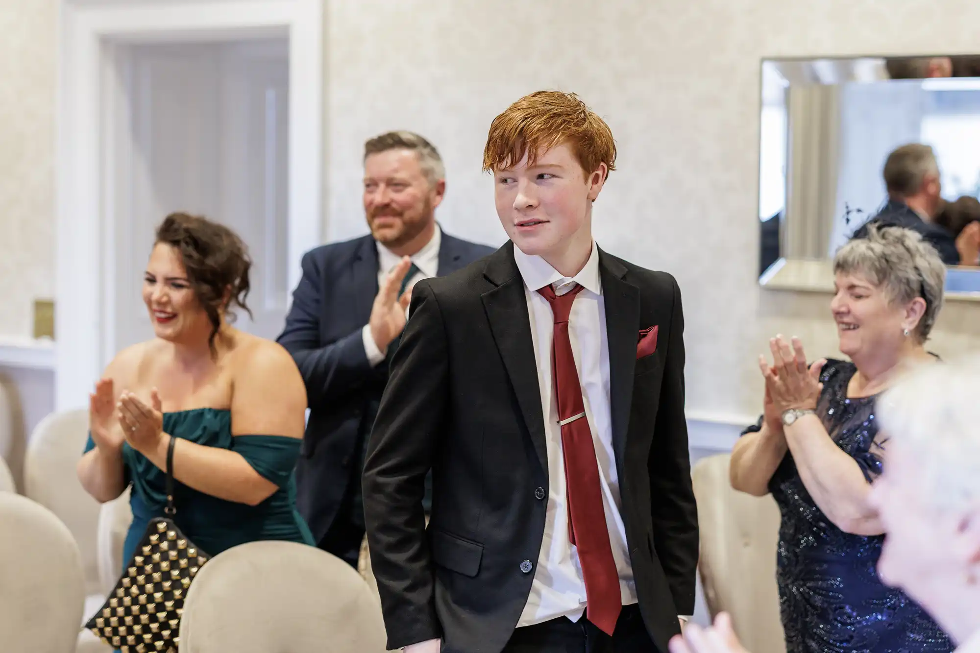 A young man with red hair in a black suit, white shirt, and red tie stands in a room as three adults, dressed formally, smile and clap for him. The atmosphere appears celebratory, with light-coloured walls and upholstered chairs in the background.