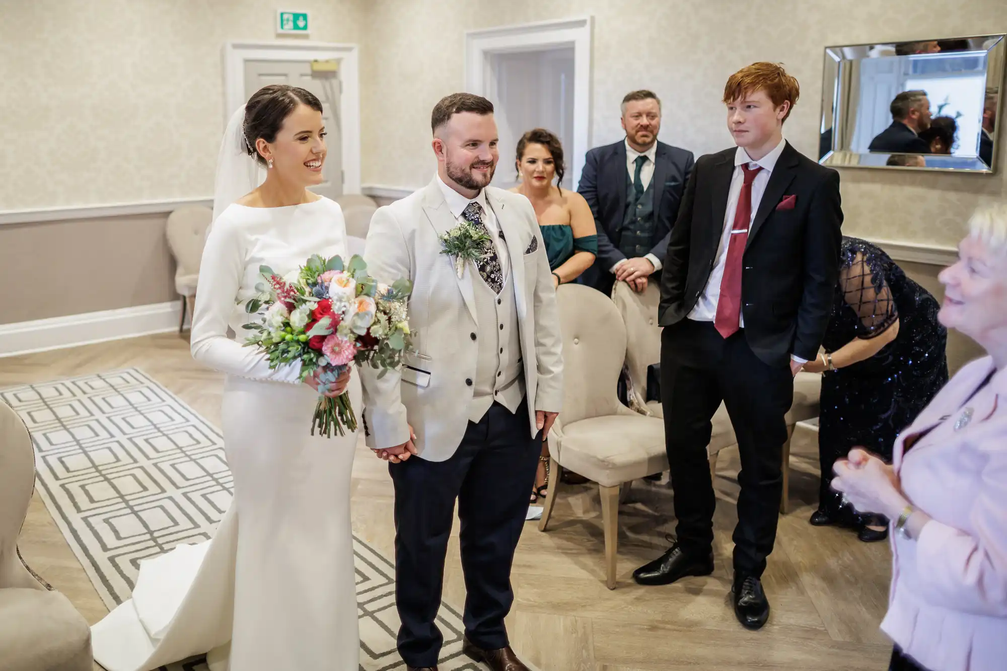 A bride in a white dress holds a colourful bouquet and stands beside the groom in a light suit. They are smiling and holding hands in a decorated room, with several guests seated and standing nearby, all dressed formally for the occasion.