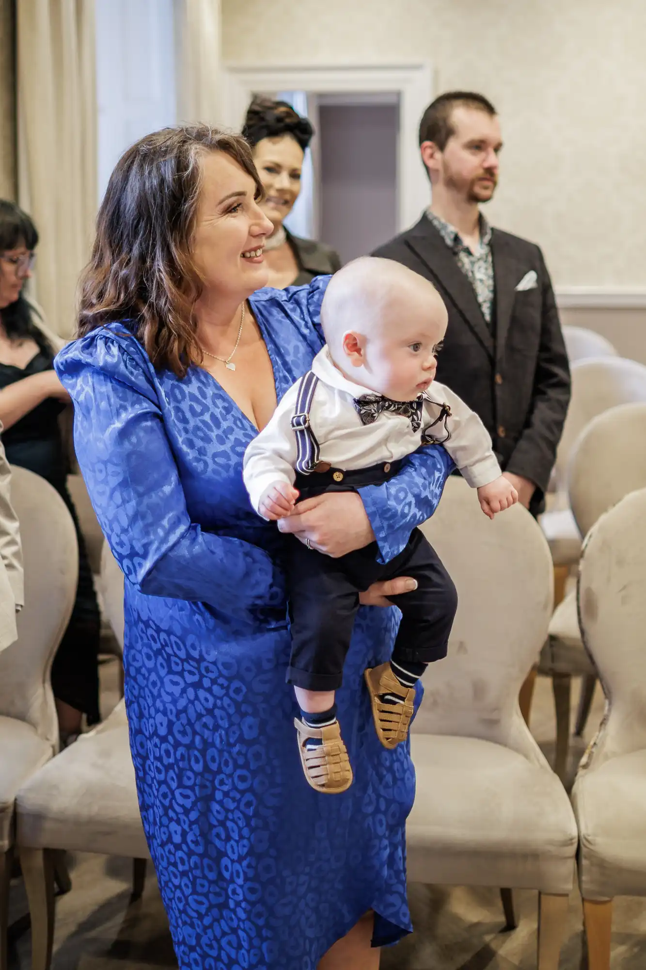 A smiling woman in a blue patterned dress holds a baby boy dressed in braces, a bow tie, and brown shoes. They are indoors, surrounded by people, with beige chairs and neutral-coloured walls in the background.