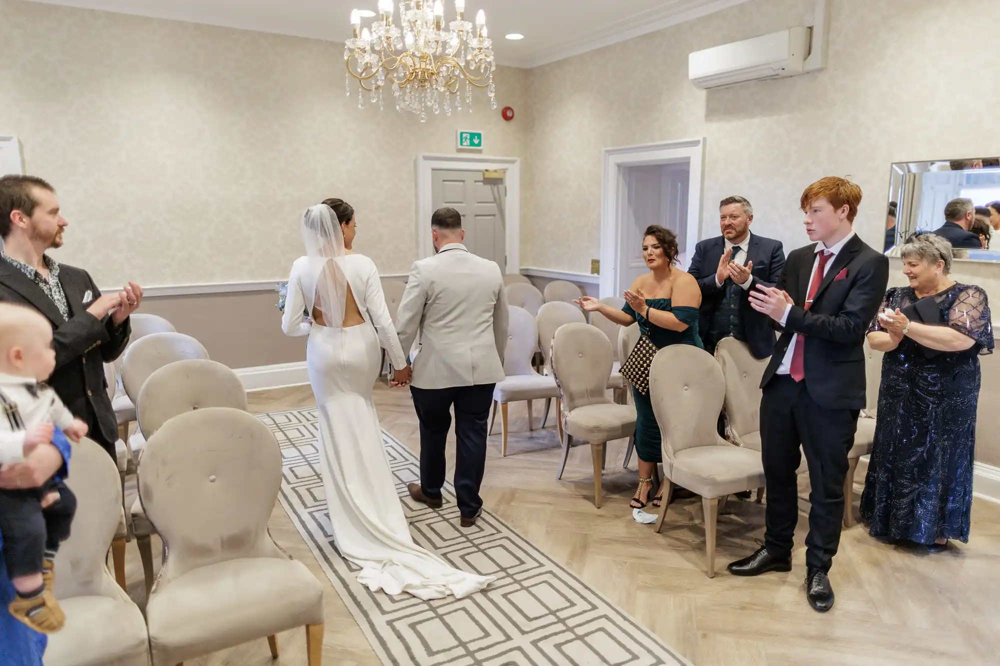 A bride in a long white dress and veil walks hand-in-hand with a groom in a light suit down an aisle in a small, elegant room. Guests standing on either side applaud, while a man holds a baby on the left. A chandelier hangs overhead.