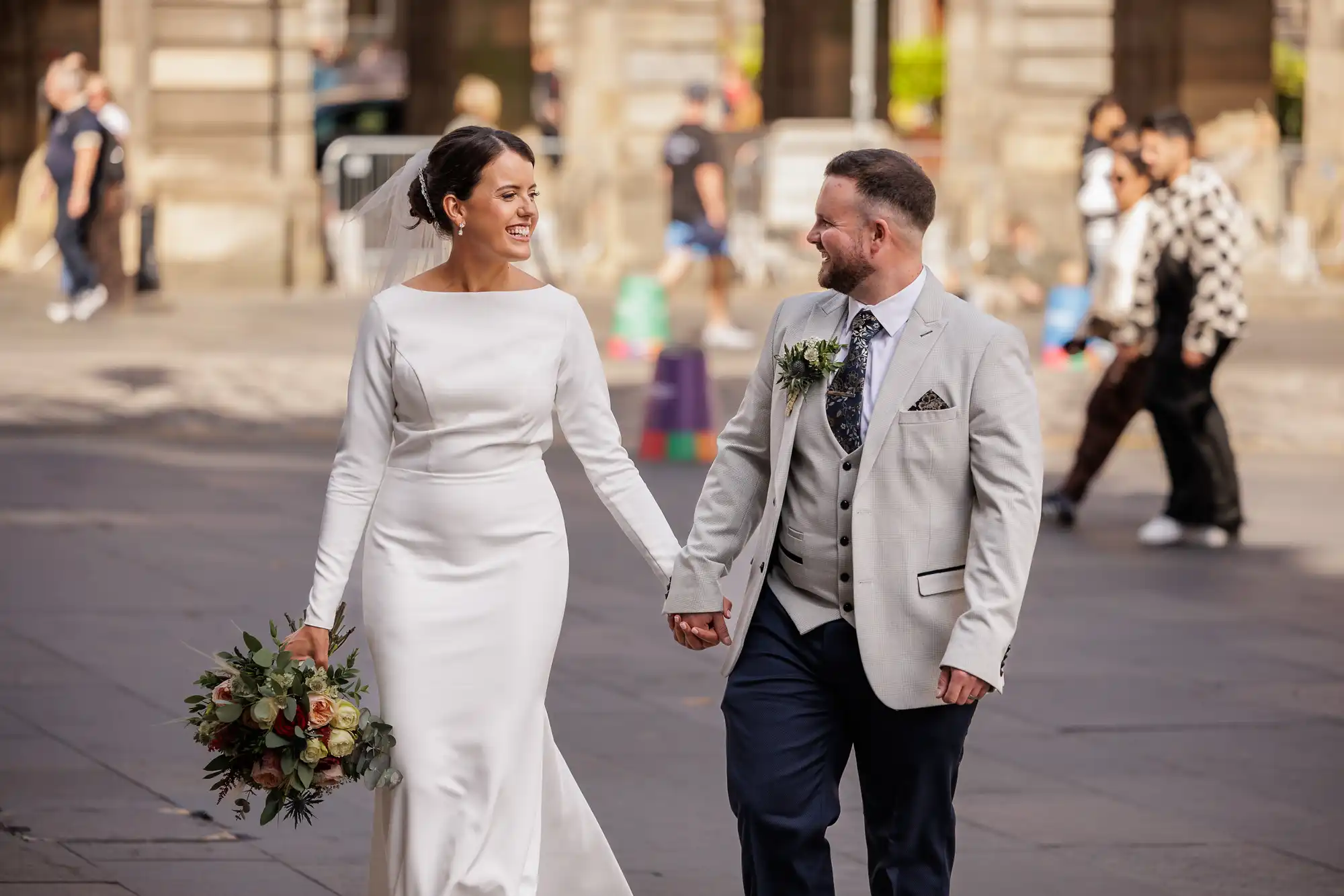 A smiling bride in a long white dress and veil holds a bunch of flowers and walks hand-in-hand with a groom in a light grey suit and patterned tie on a city street, with people blurred in the background.