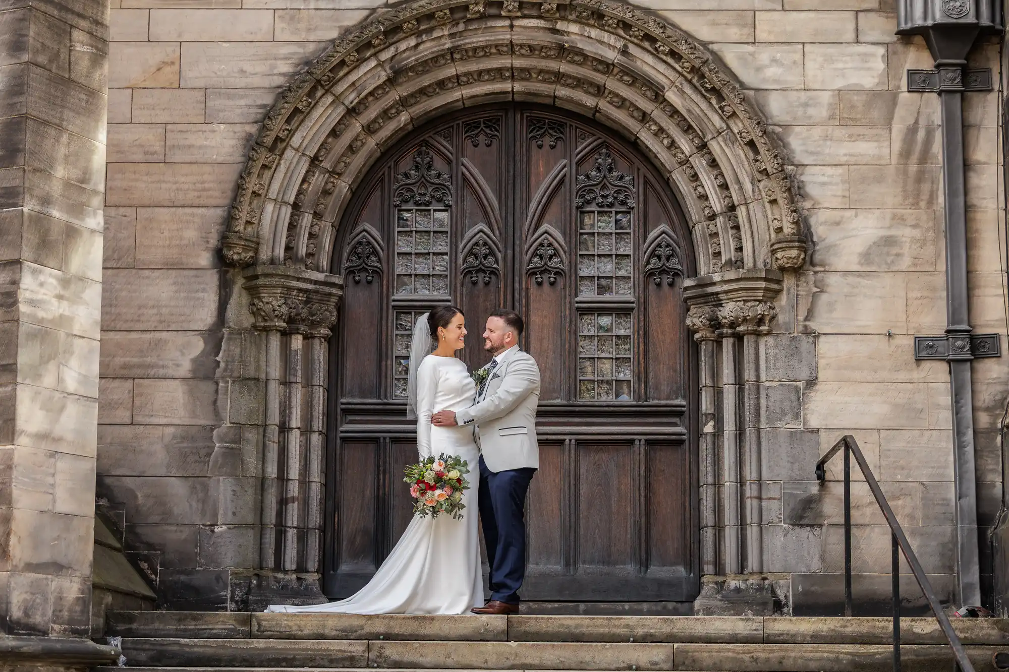 A bride in a white gown and a groom in a light grey jacket embrace on stone steps in front of a large, ornate arched wooden door with intricate carvings, set in an historic stone building. The bride holds a colourful bouquet.
