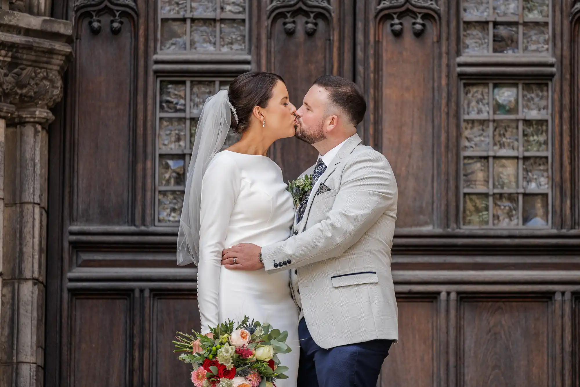 A bride in a white dress and veil holds a colourful bouquet while kissing a groom in a light jacket and dark trousers. They stand close together in front of an ornate wooden door with detailed carvings and stained glass windows.