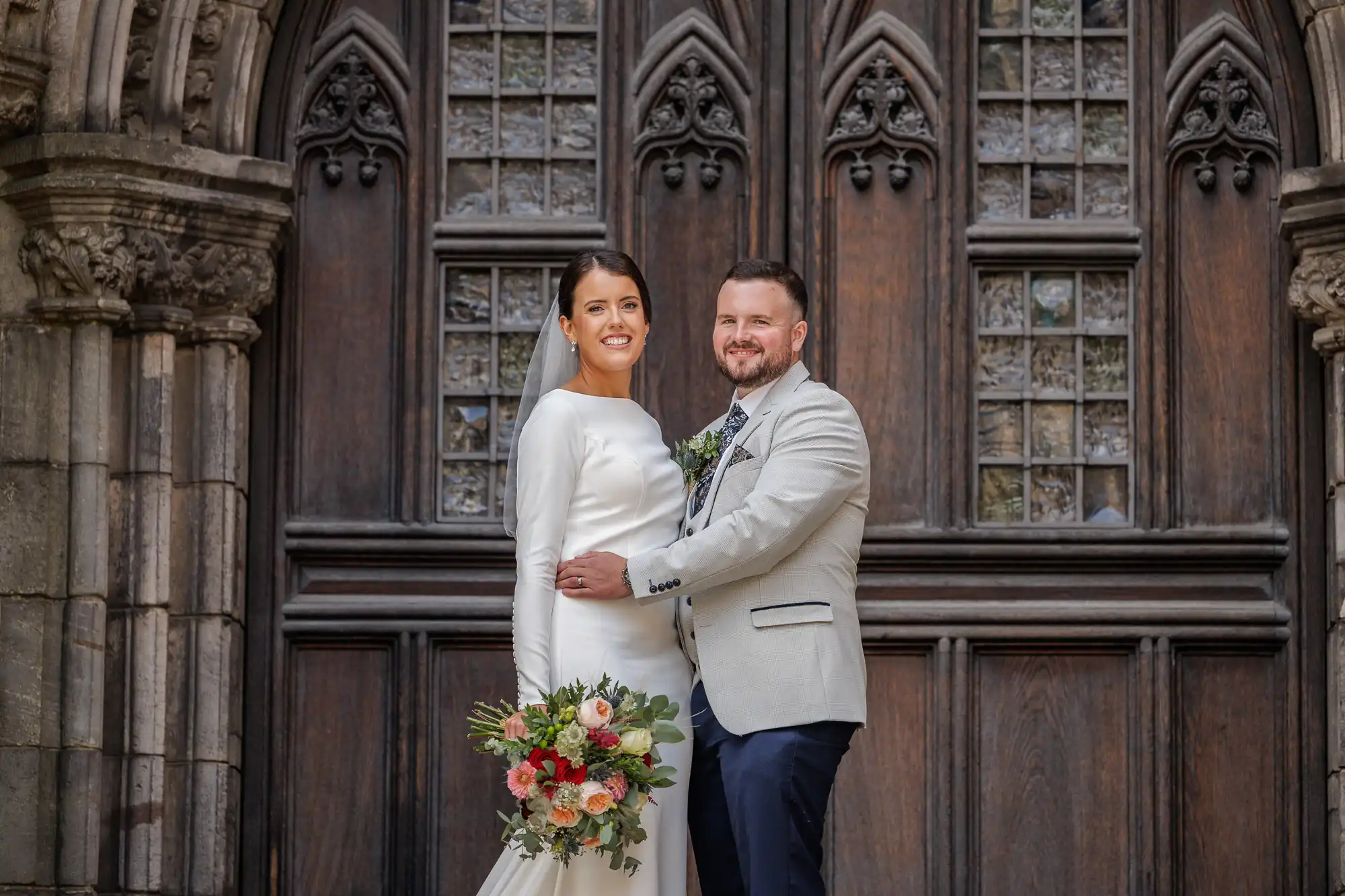 A bride in a white dress holds a bouquet of flowers while standing beside a groom in a light jacket and dark trousers. They smile and pose together in front of an ornate, wooden, gothic-style door with decorative glass panels.