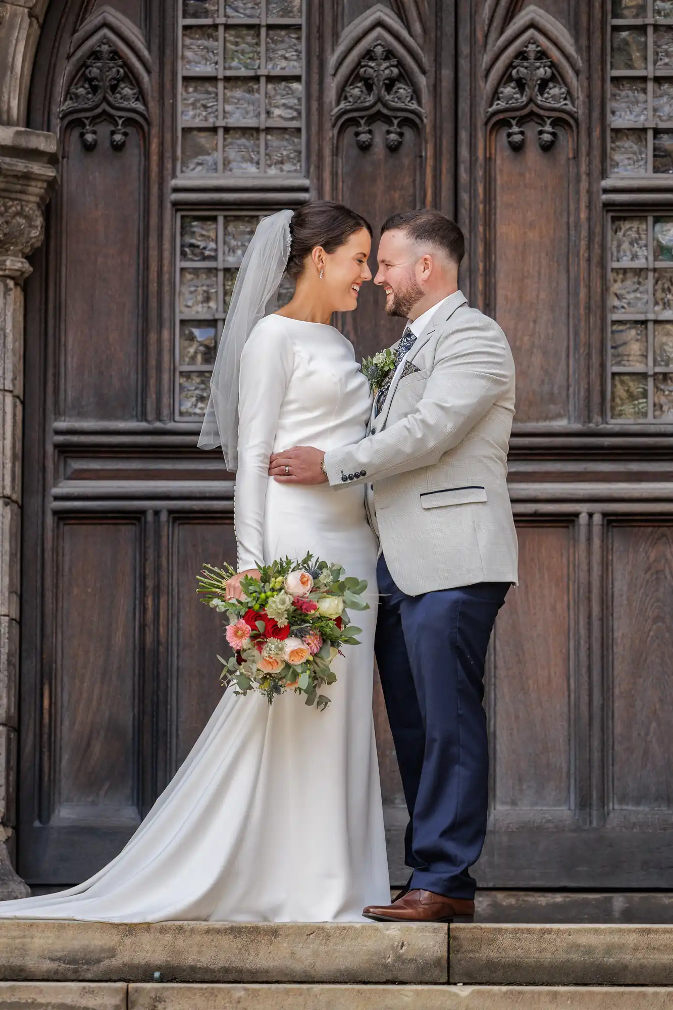 A bride in a long white gown and veil holds a bouquet of pink and peach flowers, standing close to a groom in a light grey jacket and navy trousers. They gaze at each other, smiling, in front of large wooden doors with intricate designs.