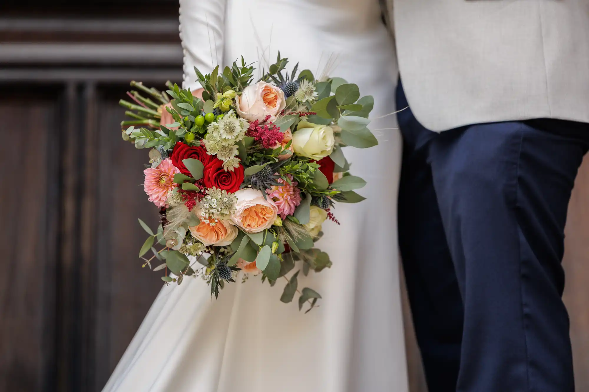 A bride in a white dress and a groom in a light jacket with dark trousers stand close together, holding a large bouquet of colourful flowers including red, pink, peach, and white roses with greenery. The background is wooden and out of focus.