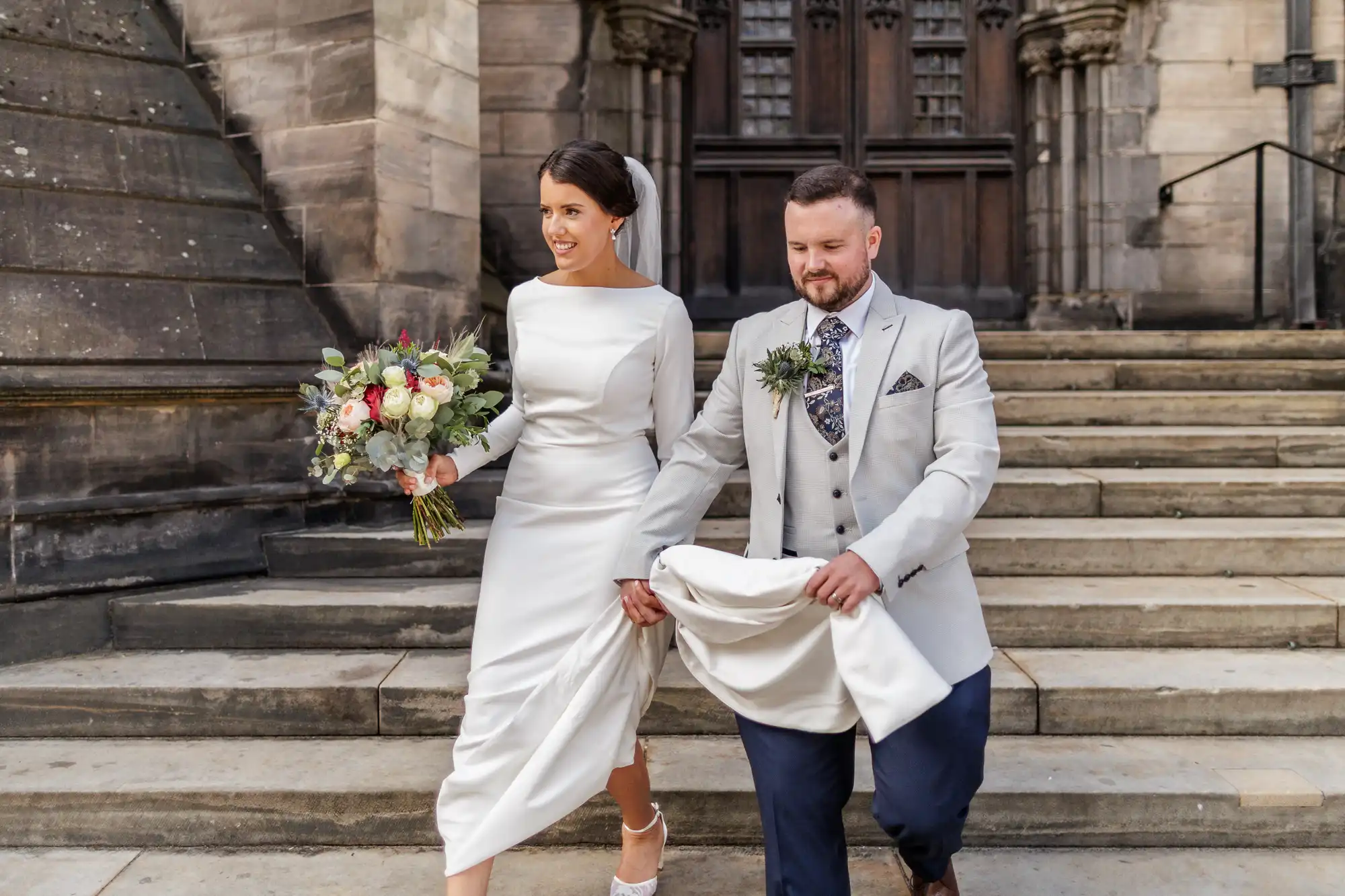A bride in a long-sleeved white dress holding a bouquet walks beside a groom in a light grey jacket and dark trousers, who lifts her train. They smile while descending stone steps outside a grand, historic building with dark wooden doors.