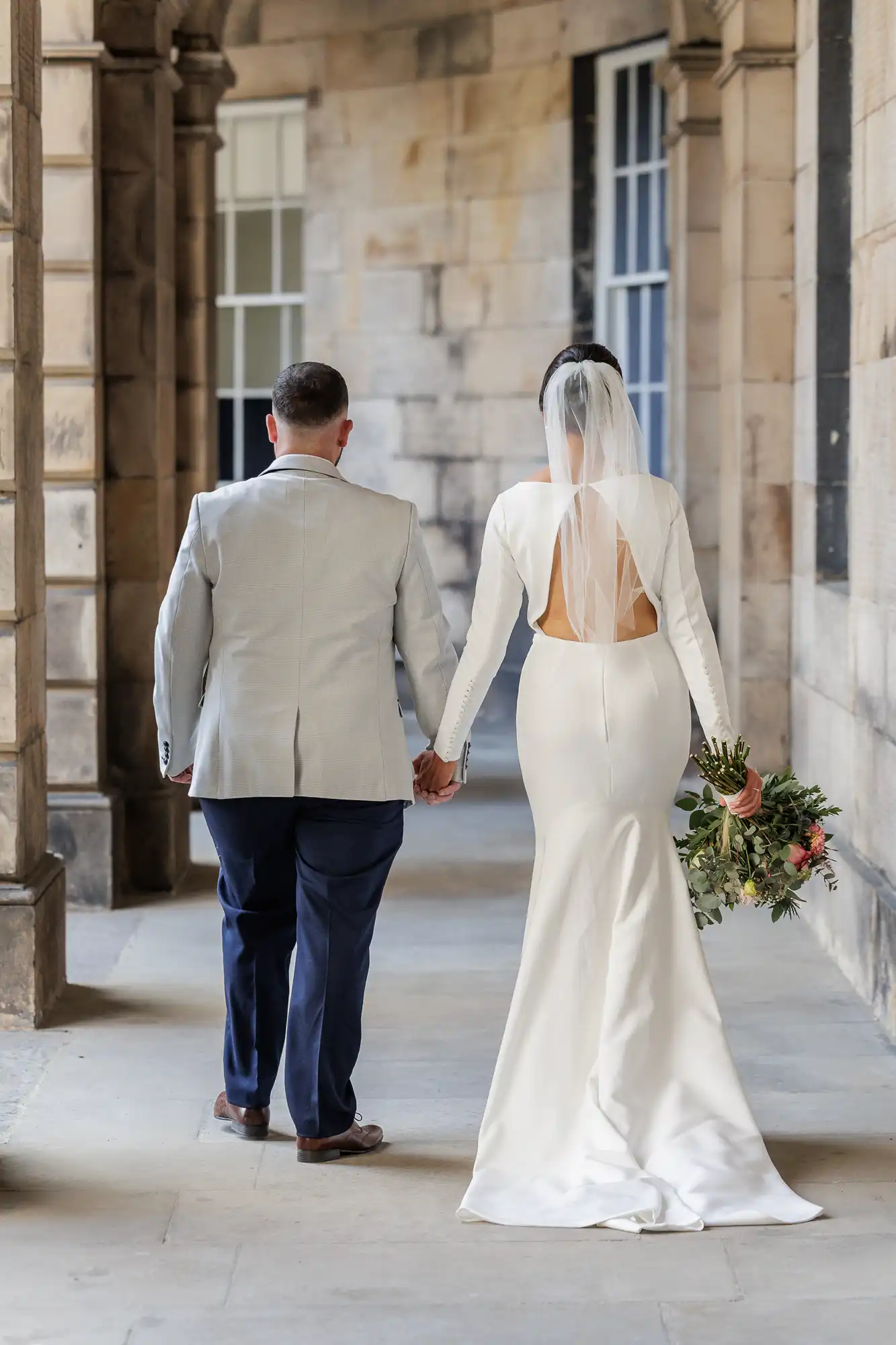 A couple walk hand in hand down a stone corridor. The bride wears a long white dress with an open back, a veil, and holds a bouquet. The groom, in a light grey jacket and dark trousers, walks beside her. Both are seen from behind.