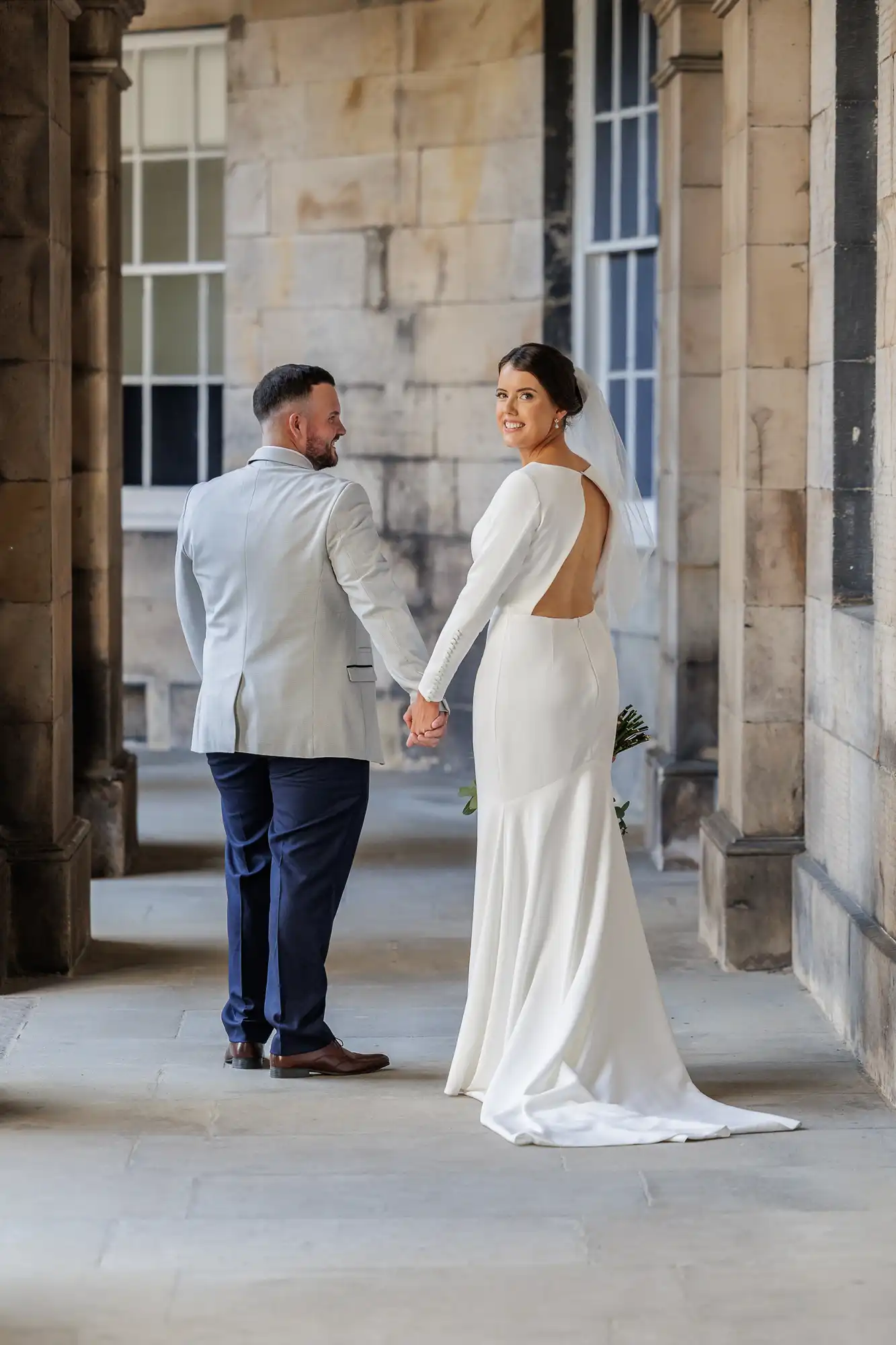 A bride in a long-sleeved, open-back white gown and veil holds hands with a groom in a light blazer and dark trousers. They walk through a stone corridor, and both glance back towards the camera, smiling. Sunlight filters through the arched walkway.
