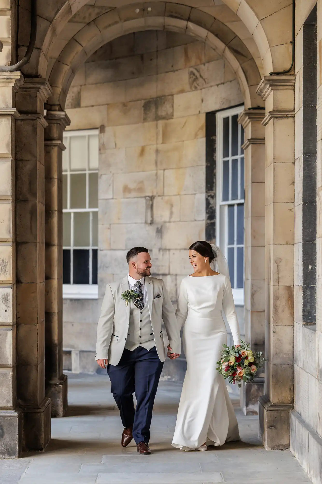 A bride in a long-sleeved white dress and veil holds a bouquet, walking hand-in-hand with a groom in a light jacket, waistcoat, and navy trousers. They smile at each other under a stone archway with tall windows behind them.