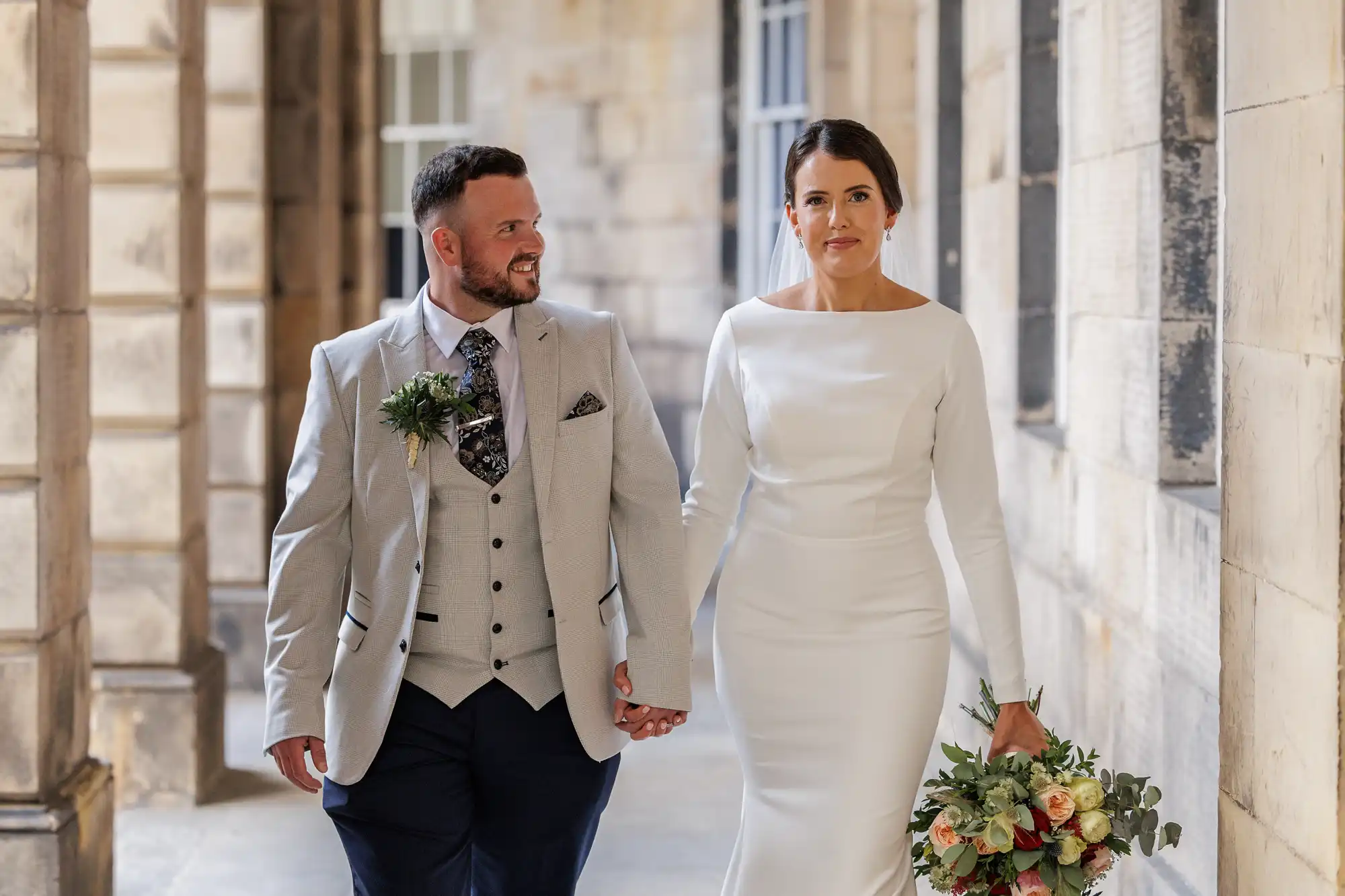 A bride in a long-sleeved white dress holds a bouquet of flowers and walks hand-in-hand with a groom in a light grey suit and patterned tie. They are outdoors, in a stone corridor, looking happy and elegant.