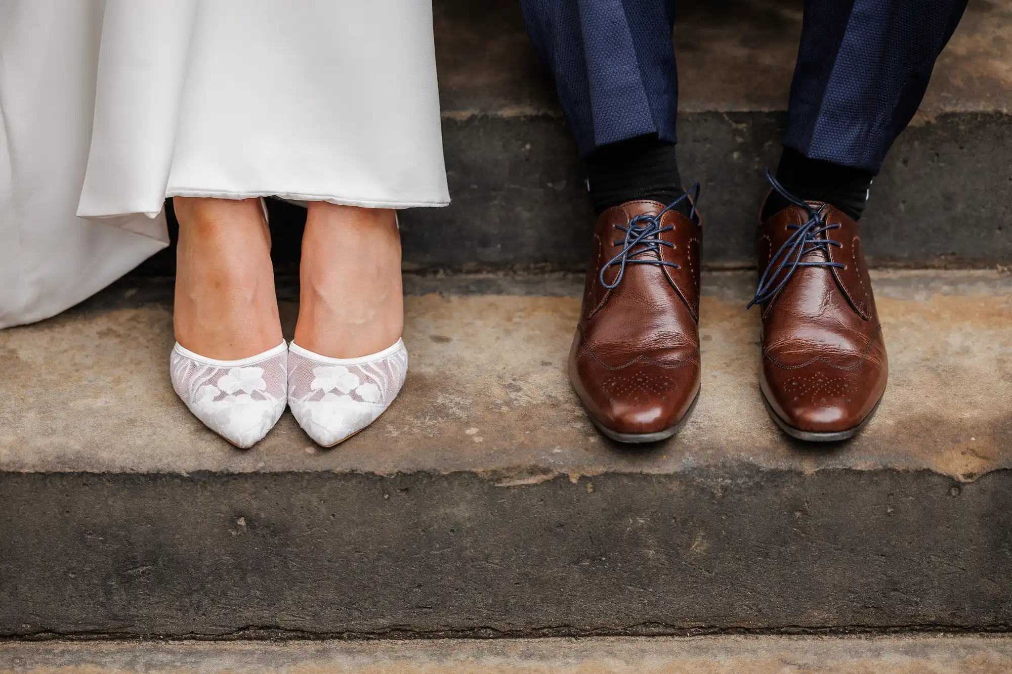 A close-up of a couple’s feet: the left side shows a person in a white dress wearing white lace pointed-toe shoes, and the right side shows a person in navy trousers wearing brown leather dress shoes with blue laces. Both are sitting on stone steps.