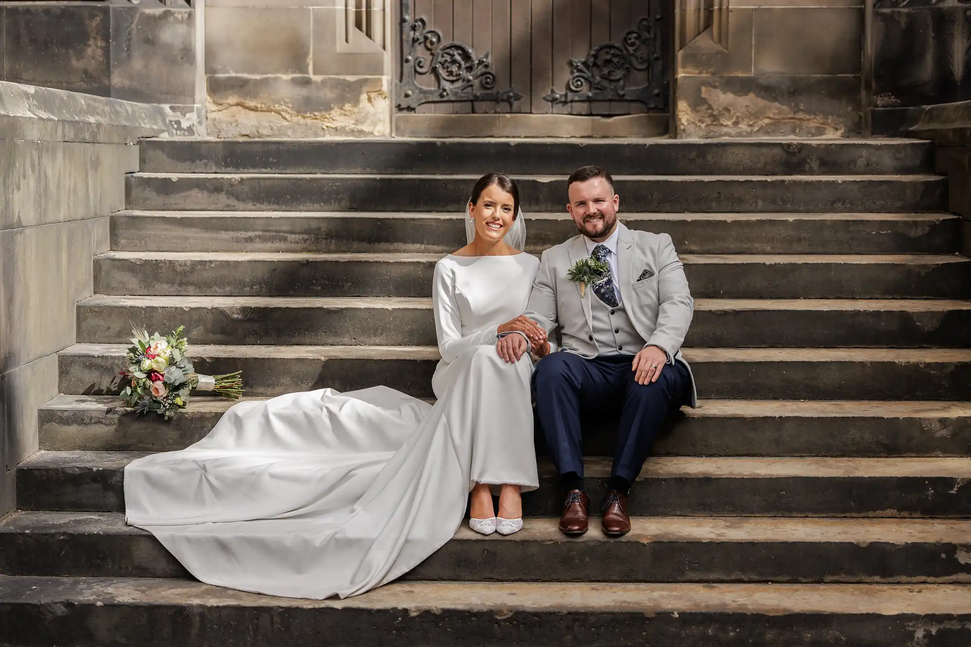 A bride in a long-sleeved white gown and a groom in a light grey suit sit smiling on stone steps. A bouquet of flowers rests to the bride’s left. The background features a worn stone wall and ornate iron gate.