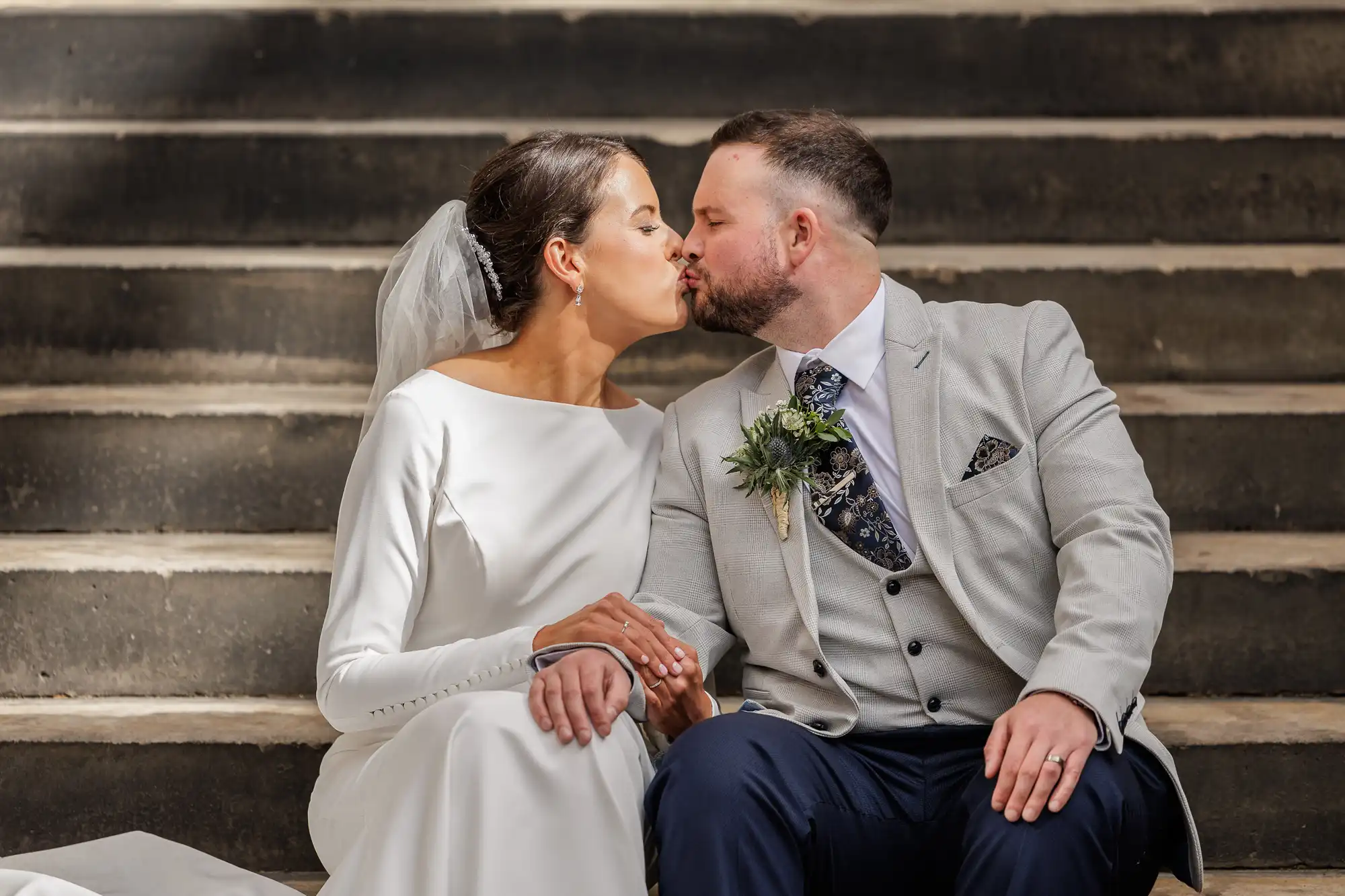 A bride in a long-sleeved white dress and veil kisses a groom in a light grey suit with a buttonhole as they sit holding hands on outdoor stone steps, sunlight highlighting their faces and expressions of happiness.